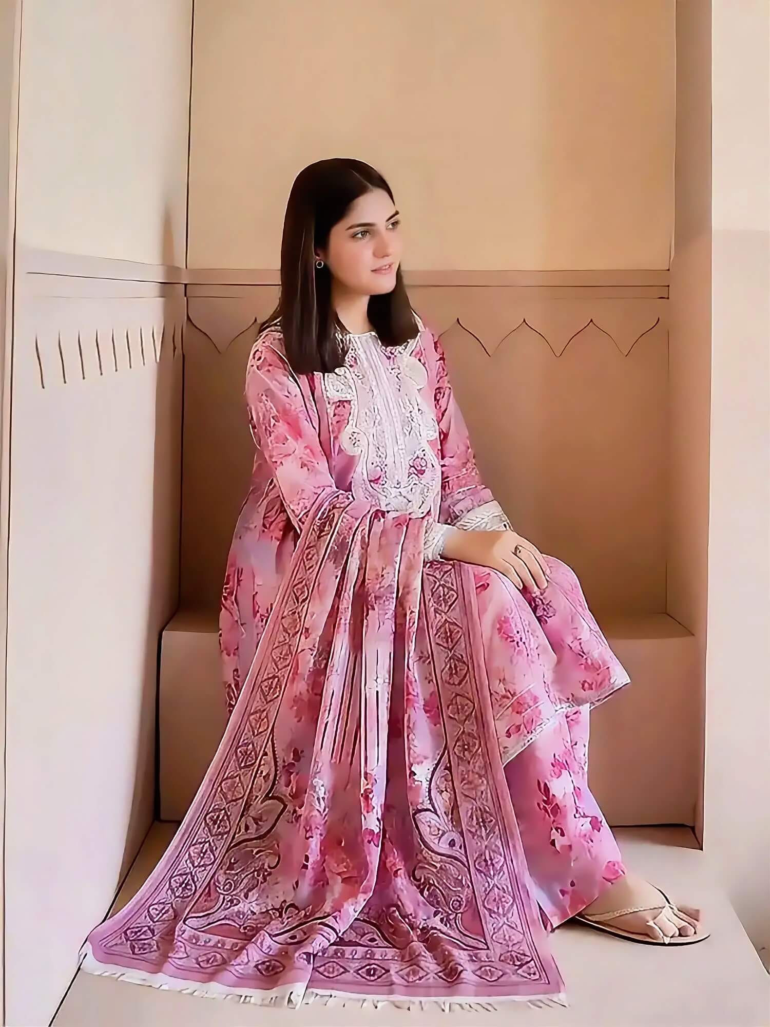A woman wearing a pink printed traditional outfit with a matching dupatta, seated on a beige bench in a minimalist indoor setting.