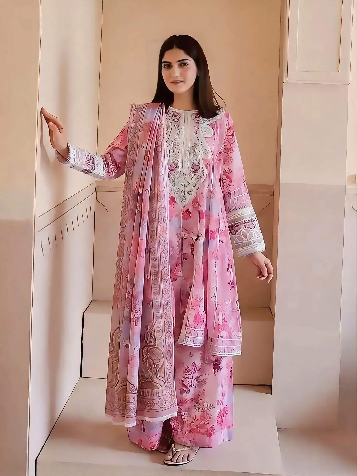 A woman modeling a pink floral traditional outfit with a matching printed dupatta, standing in a beige indoor setting.