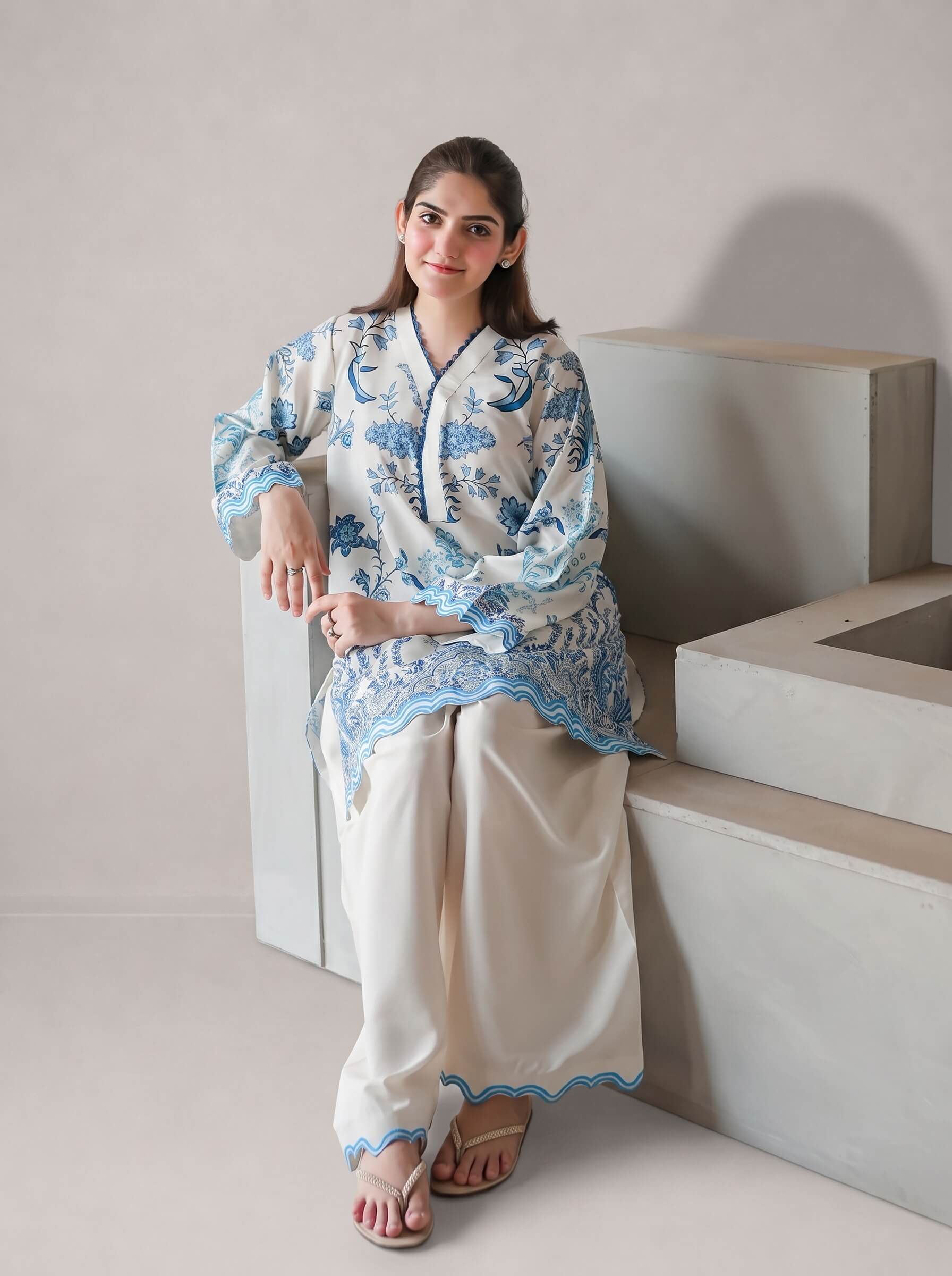 A woman models a traditional South Asian outfit consisting of a white kurta with blue floral embroidery and wide-leg cream trousers, sitting on minimalist concrete steps against a neutral beige wall.