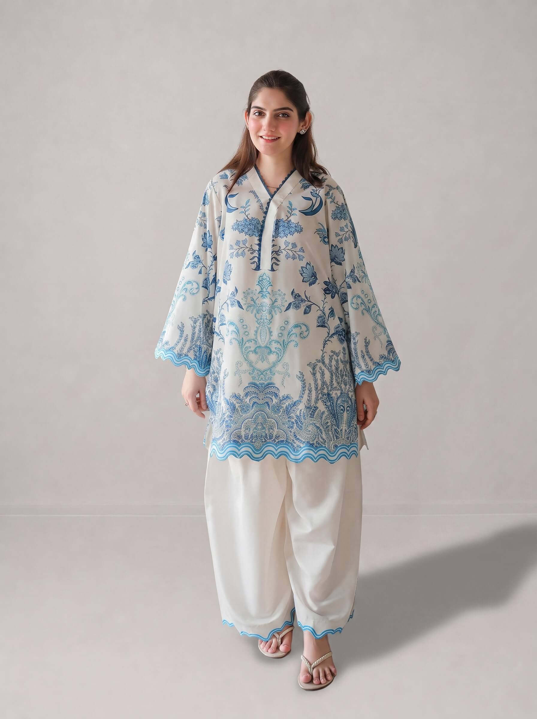 A woman modeling a traditional South Asian outfit consisting of a cream-colored kurta with blue floral embroidery and wide-leg cream trousers, set against a plain light gray studio background.
