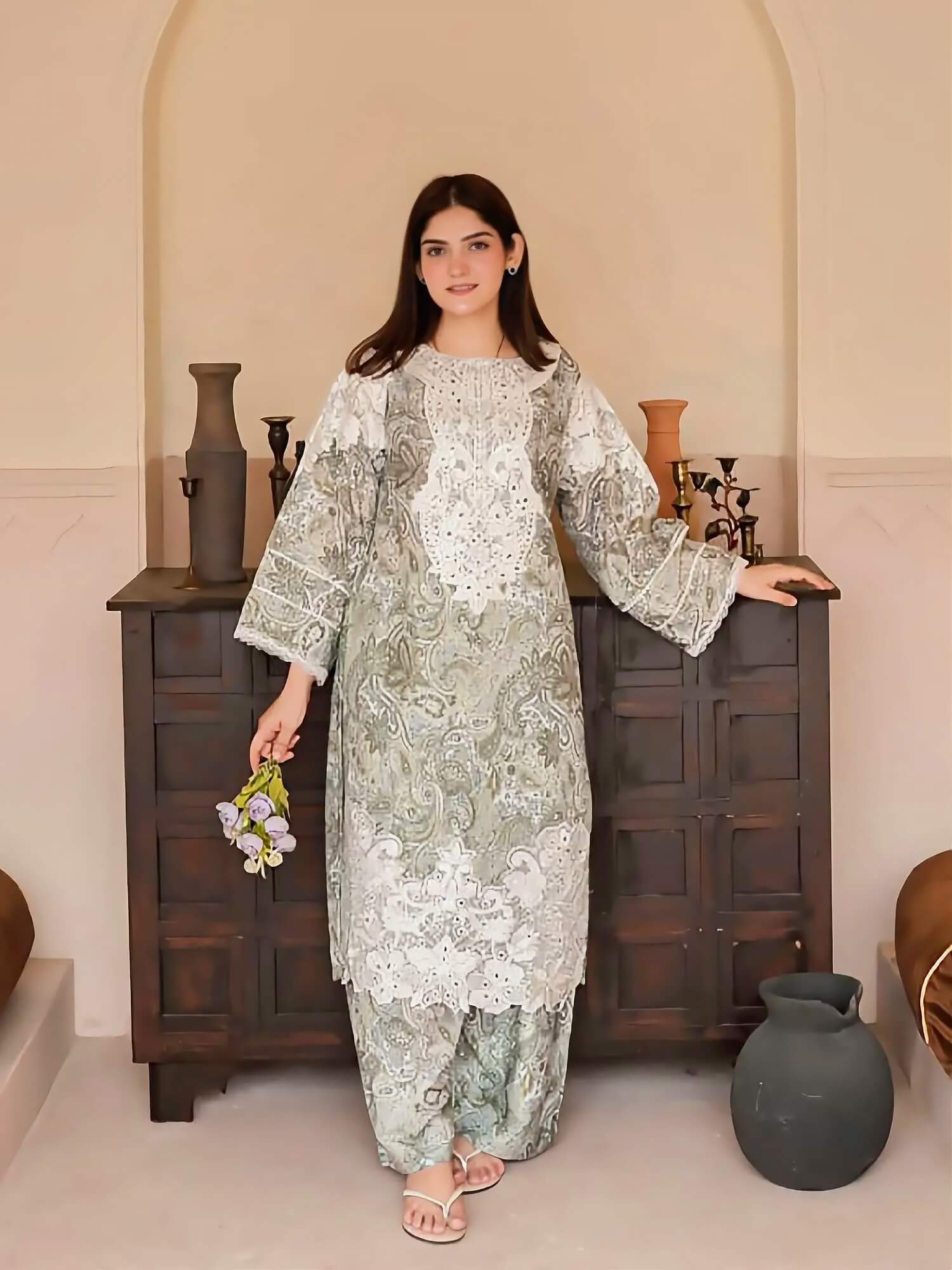 A woman models a three-piece traditional outfit with a sage green and white paisley pattern, standing in front of a dark wooden cabinet in a neutral indoor setting.