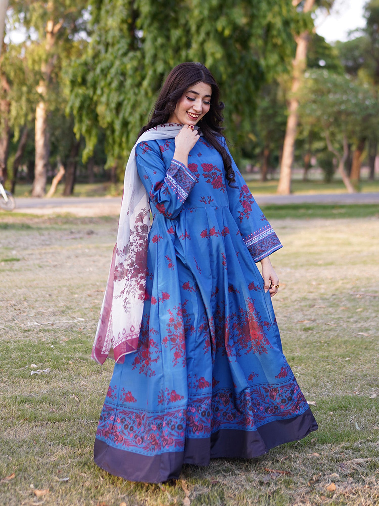 A woman wearing a blue traditional long dress with red floral prints and a matching dupatta, standing outdoors on a grassy park path with trees in the background.