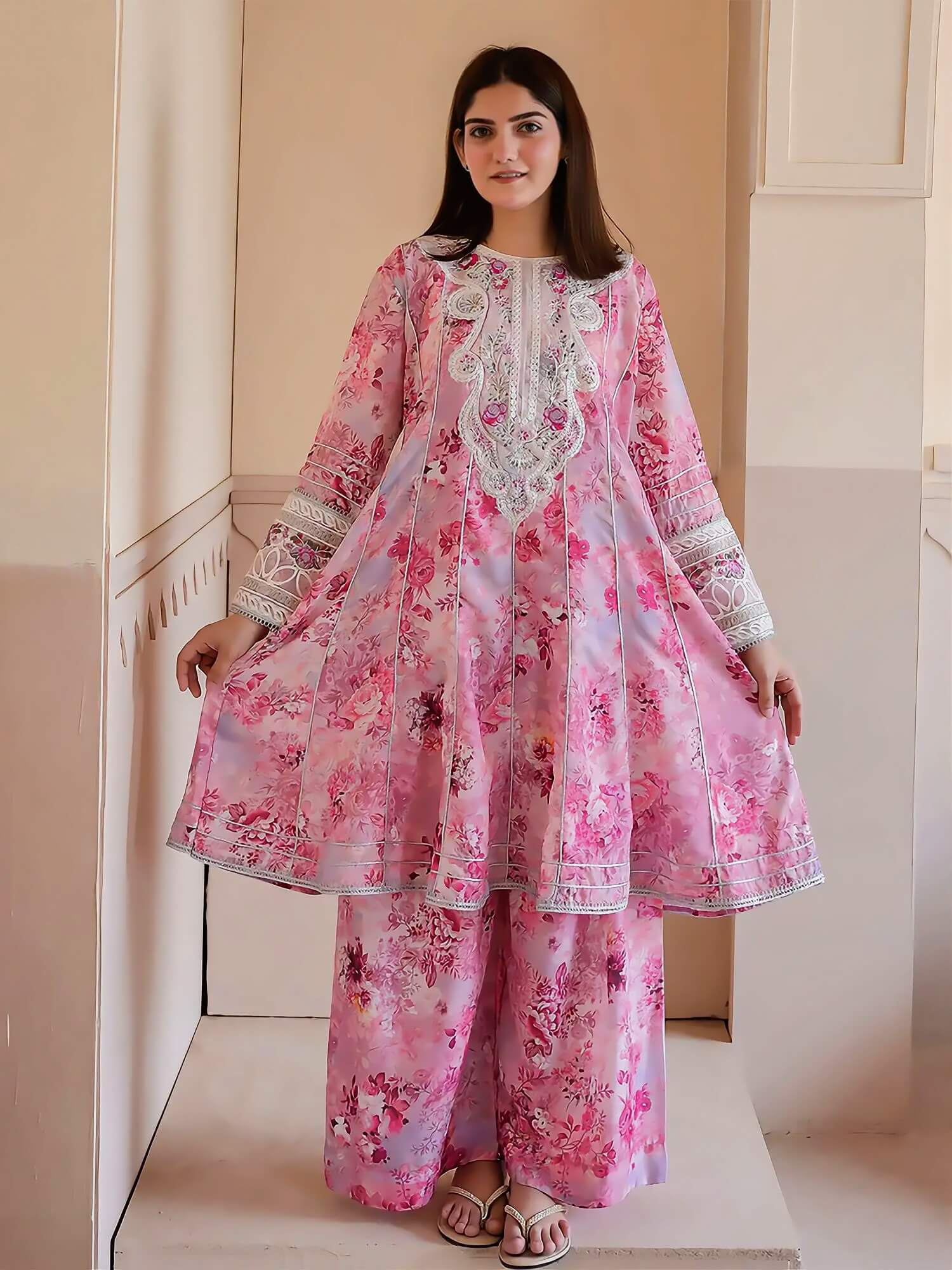 A woman modeling a pink floral traditional outfit with white embroidered details, standing in an indoor setting with beige walls.
