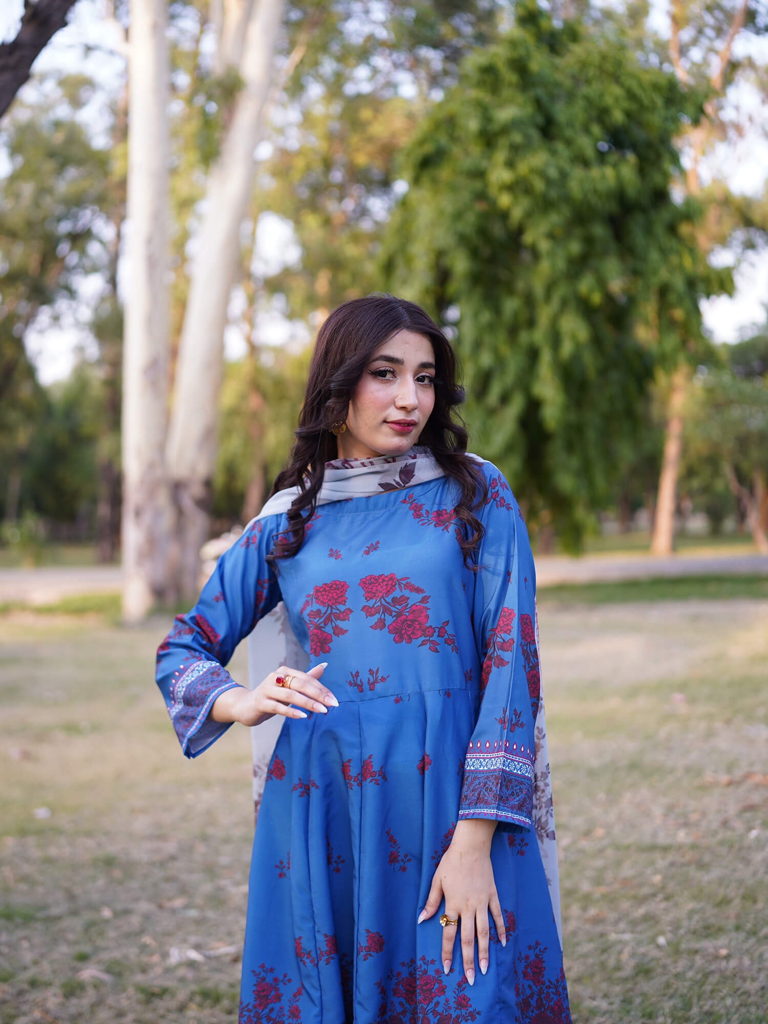 A woman wearing a blue traditional dress with red floral patterns and a gray patterned scarf, standing in a grassy park with trees in the background.