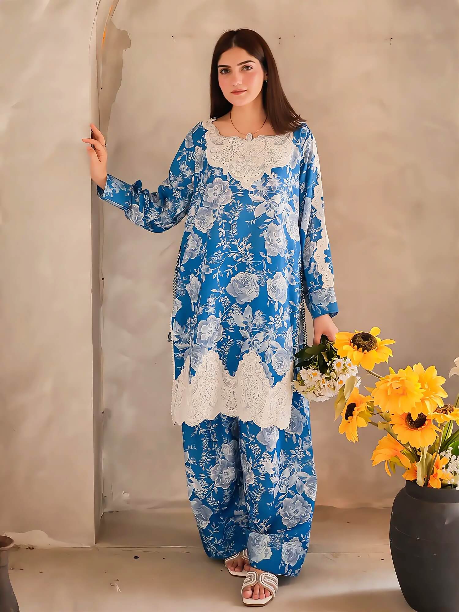 A woman modeling a blue and white floral printed kurta set with white lace trim, standing in a rustic beige room next to a black vase of yellow sunflowers.