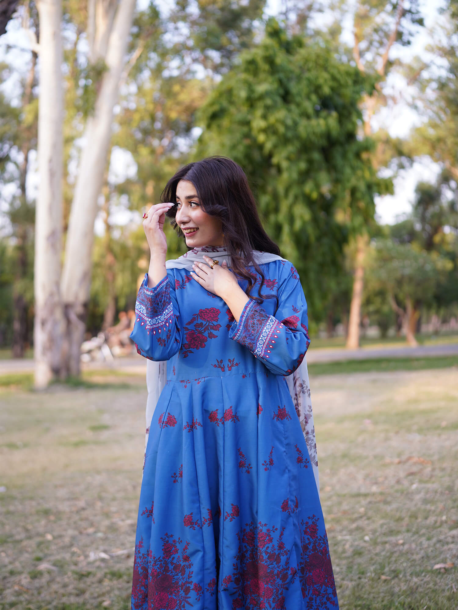 A woman wearing a blue traditional long dress with red floral patterns and a matching scarf, standing in a grassy park with trees in the background.
