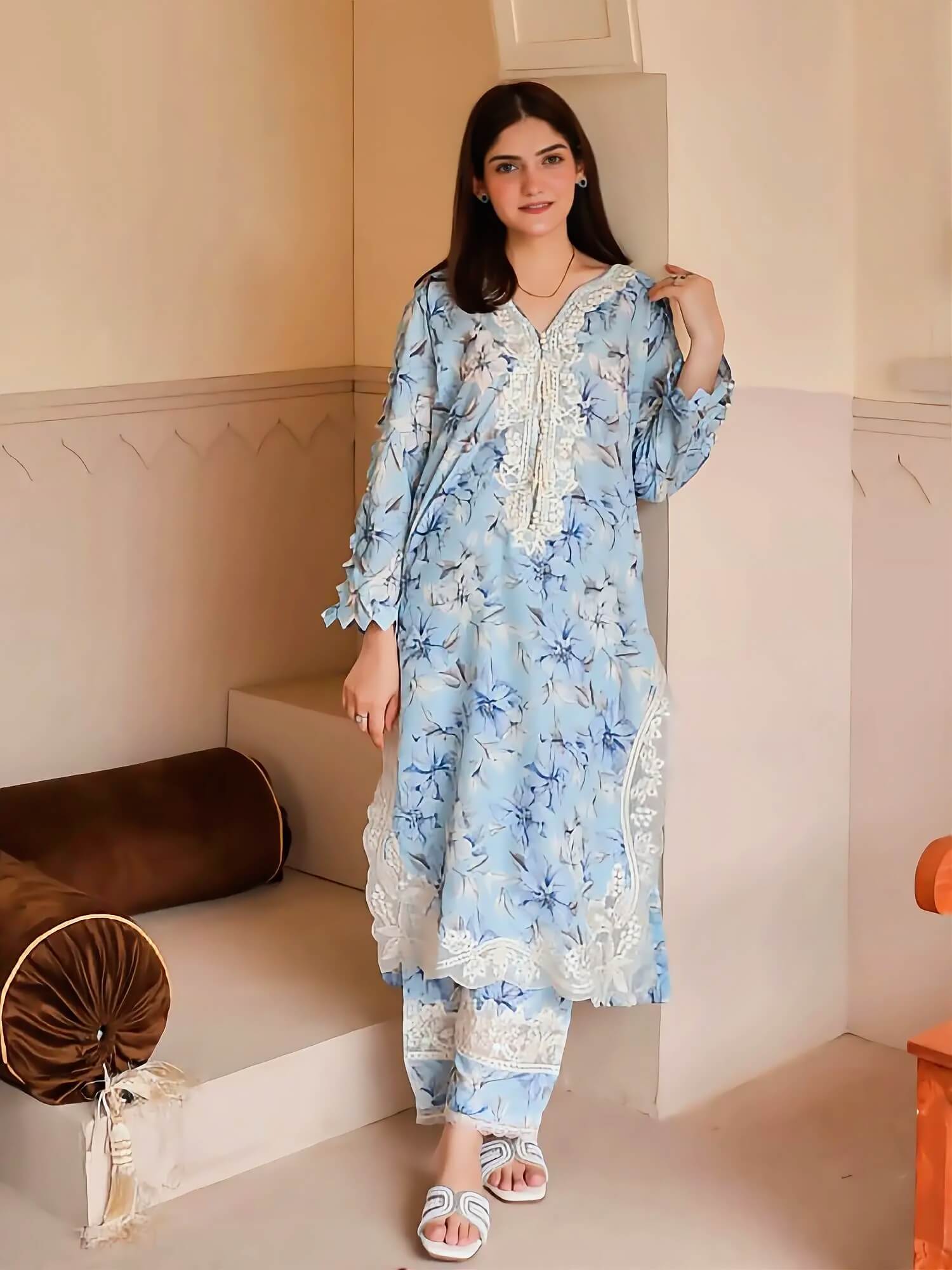 A woman models a light blue and white floral printed three-piece kurta set with white lace details, standing in a beige-toned indoor setting.
