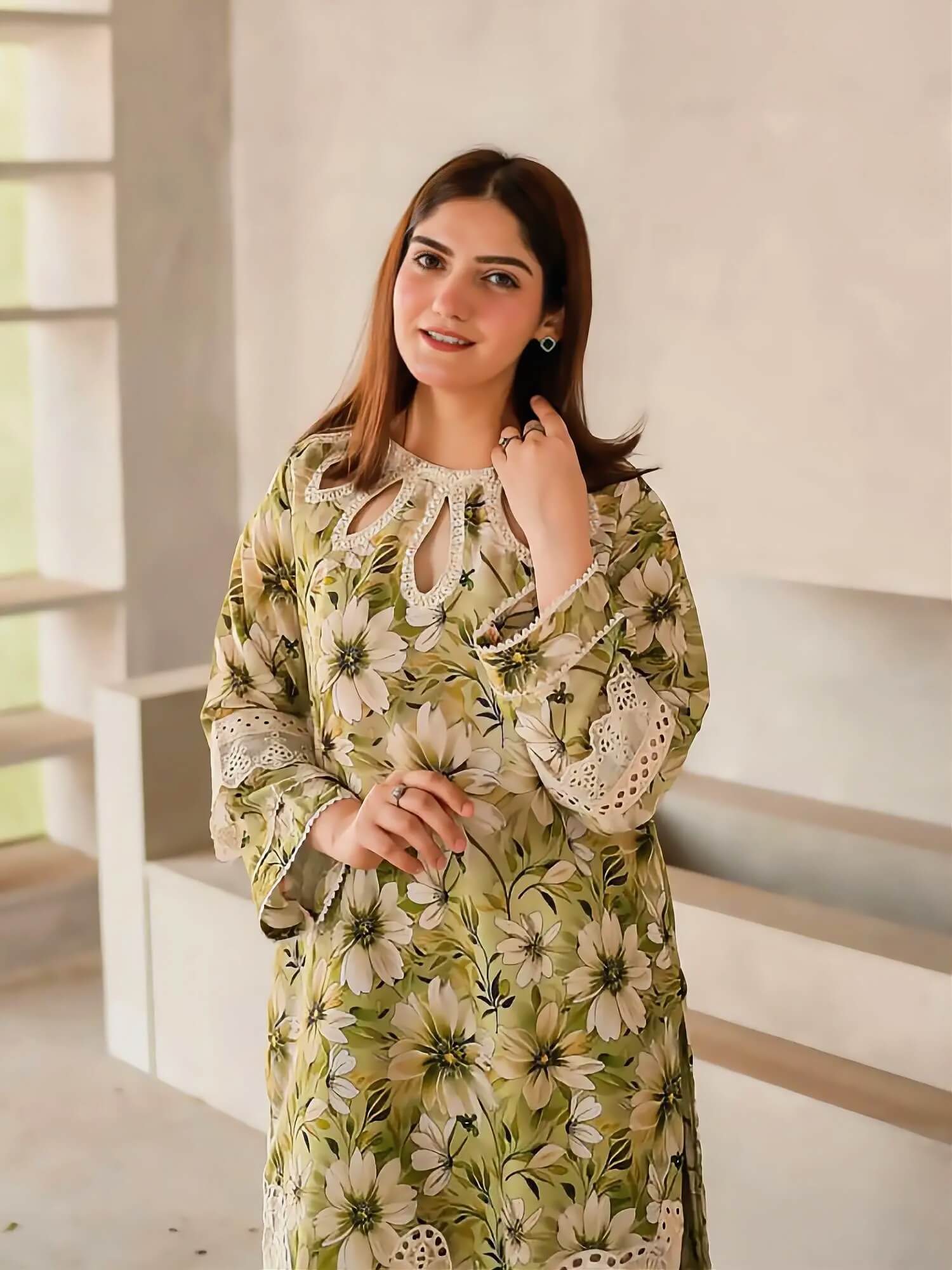 A woman wearing a long-sleeved olive green and beige floral print tunic dress with white lace details, standing in a modern, minimalist interior with light-colored steps.