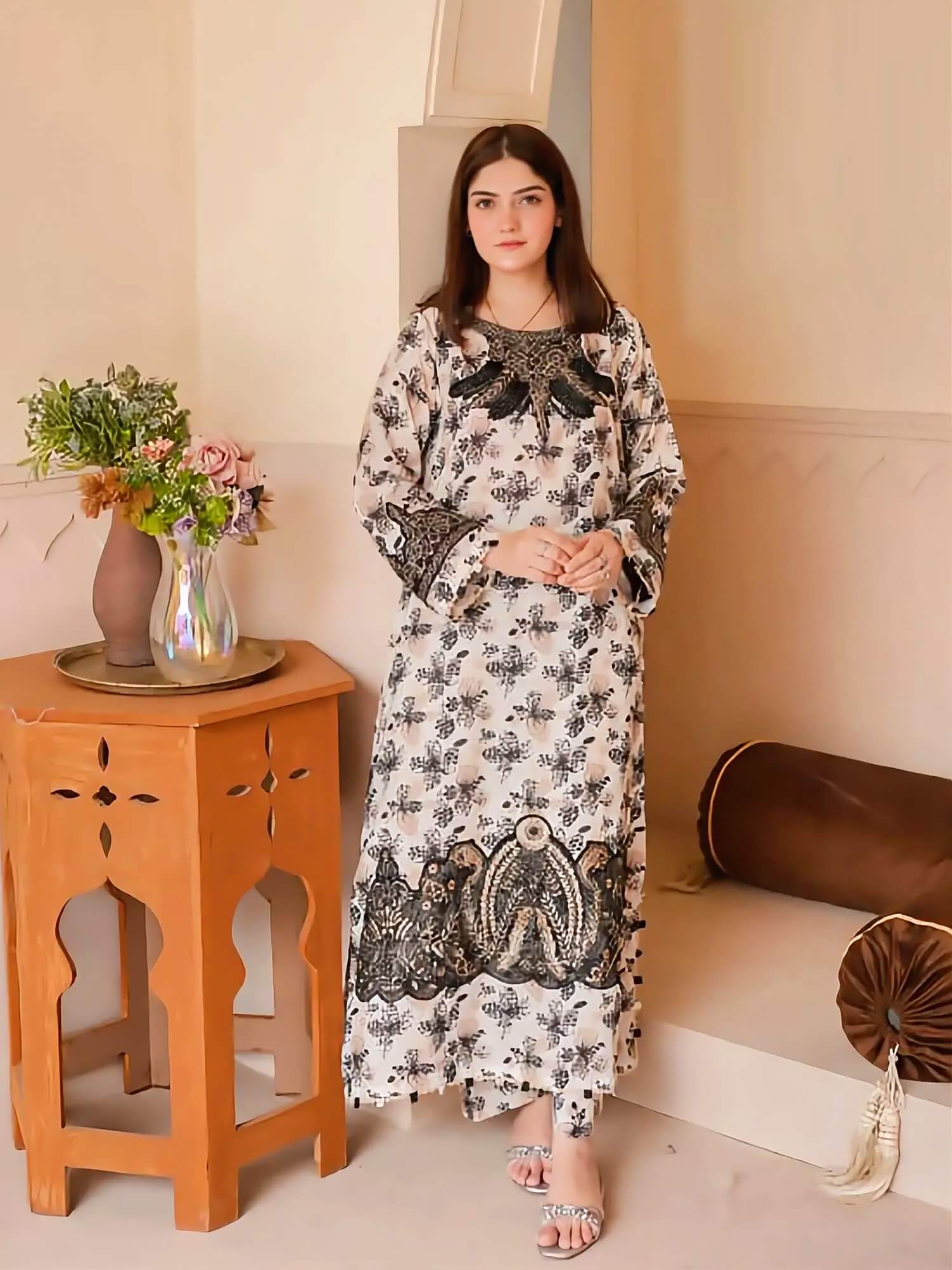A woman models a long-sleeved, floor-length kurta dress with a black and white floral pattern and black embroidery, standing in a warm-toned indoor setting next to a wooden side table.