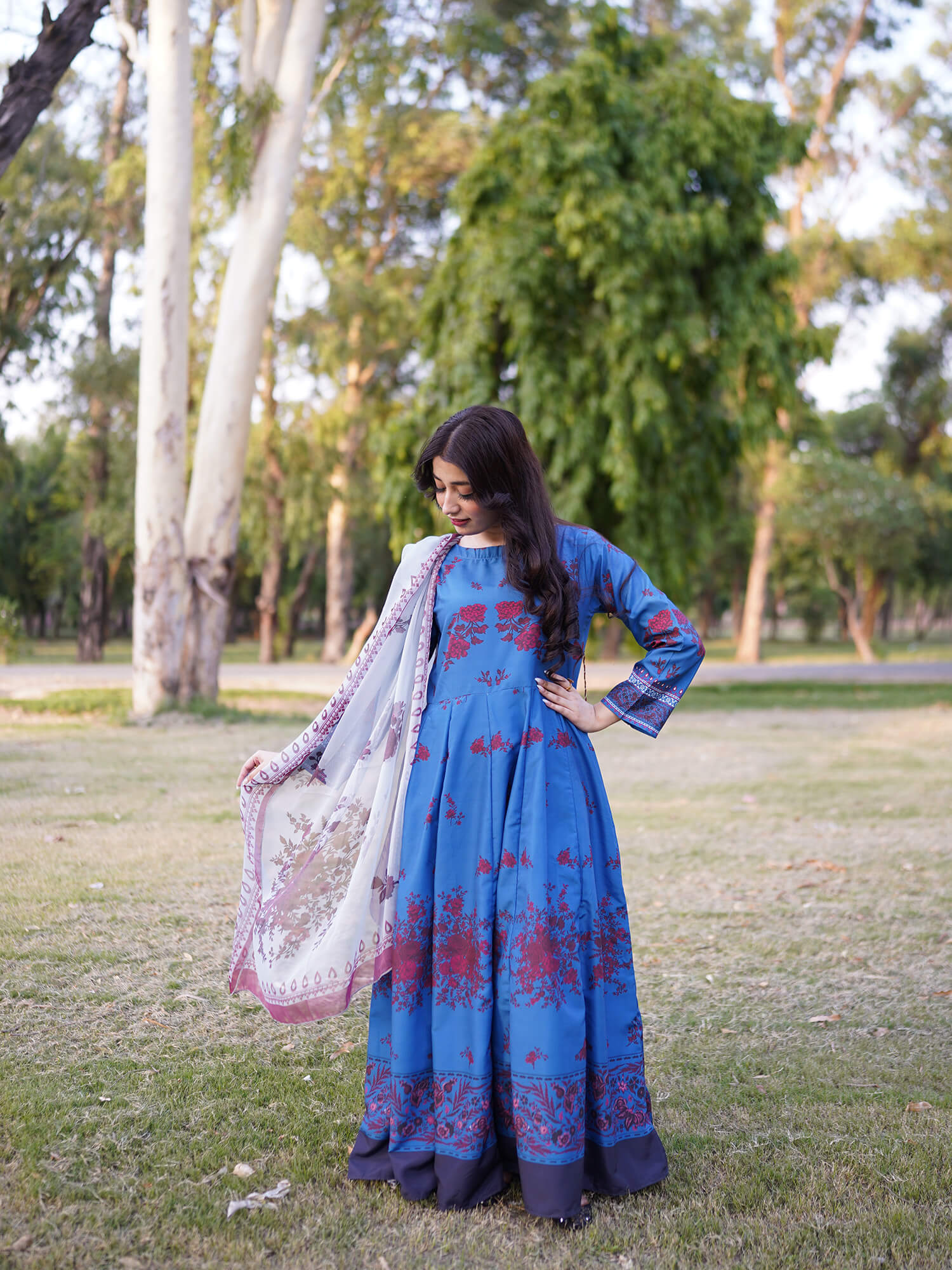 A woman stands outdoors on a grassy park lawn wearing a blue long-sleeved traditional dress with red floral embroidery and holding a white dupatta with pink and blue patterns.