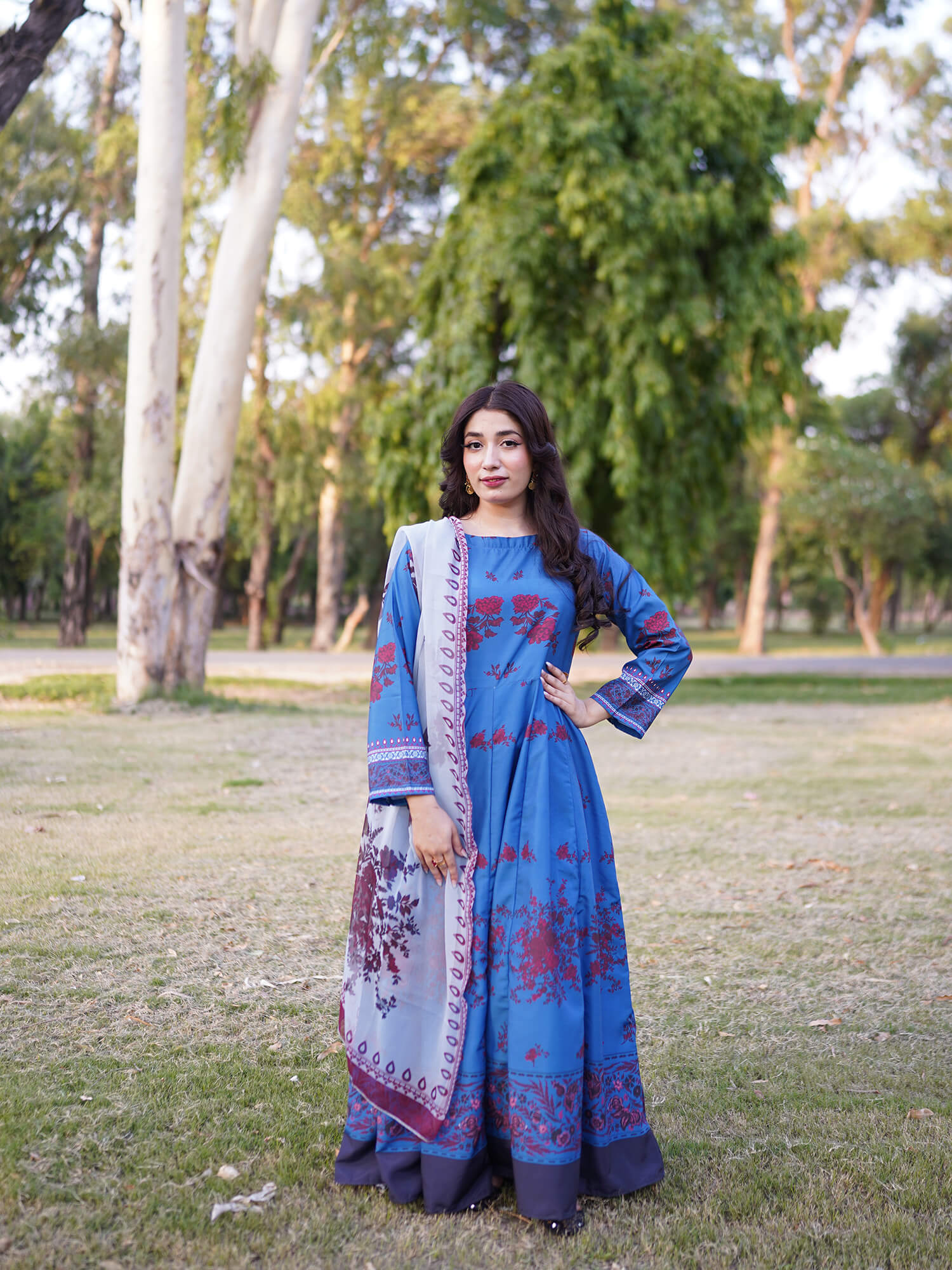A woman stands outdoors in a park wearing a blue traditional long dress with red floral patterns and a matching light blue dupatta with purple borders.
