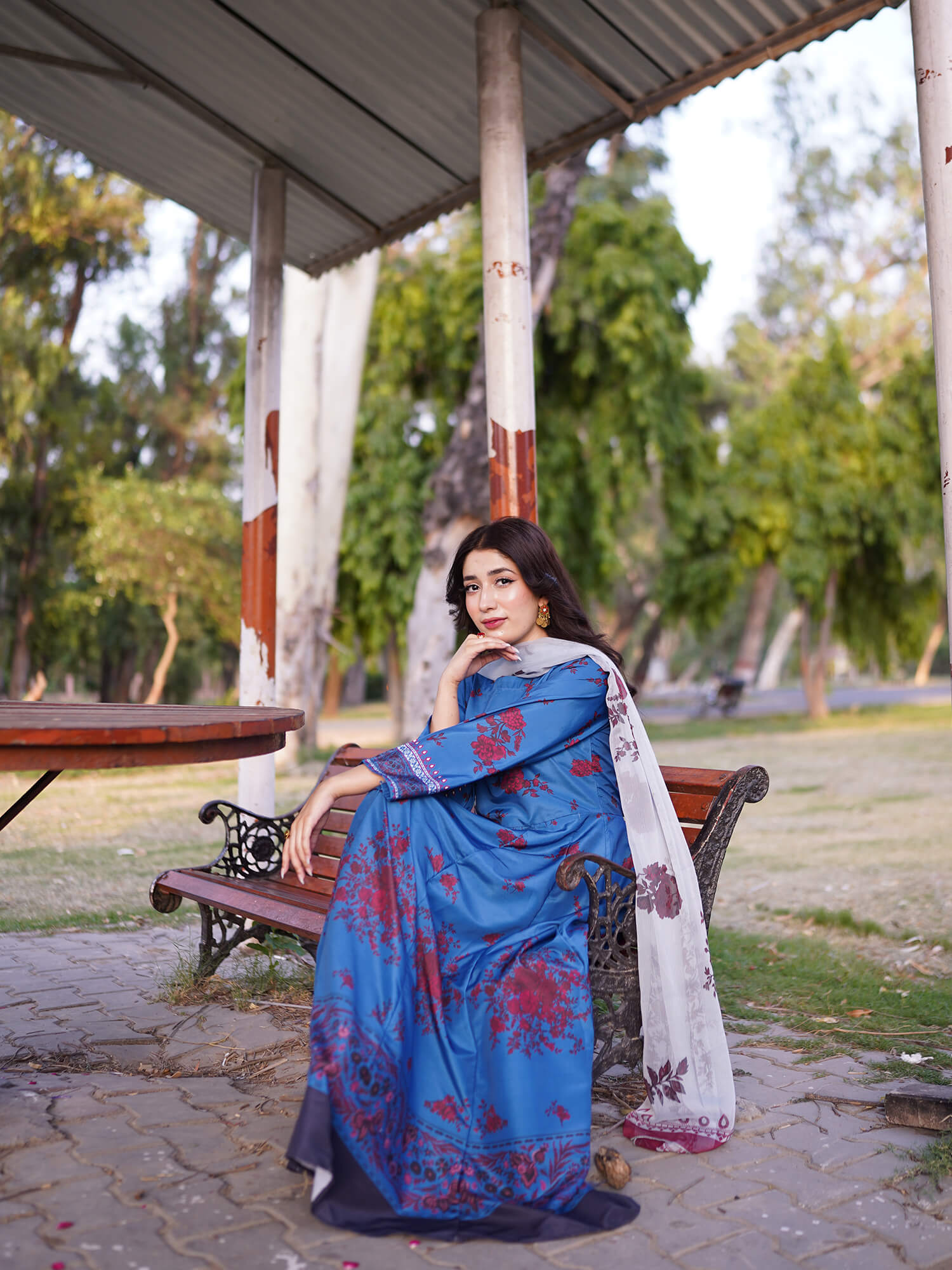 A woman wearing a blue floral printed salwar suit with a white dupatta sits on a wooden bench under a covered outdoor pavilion.