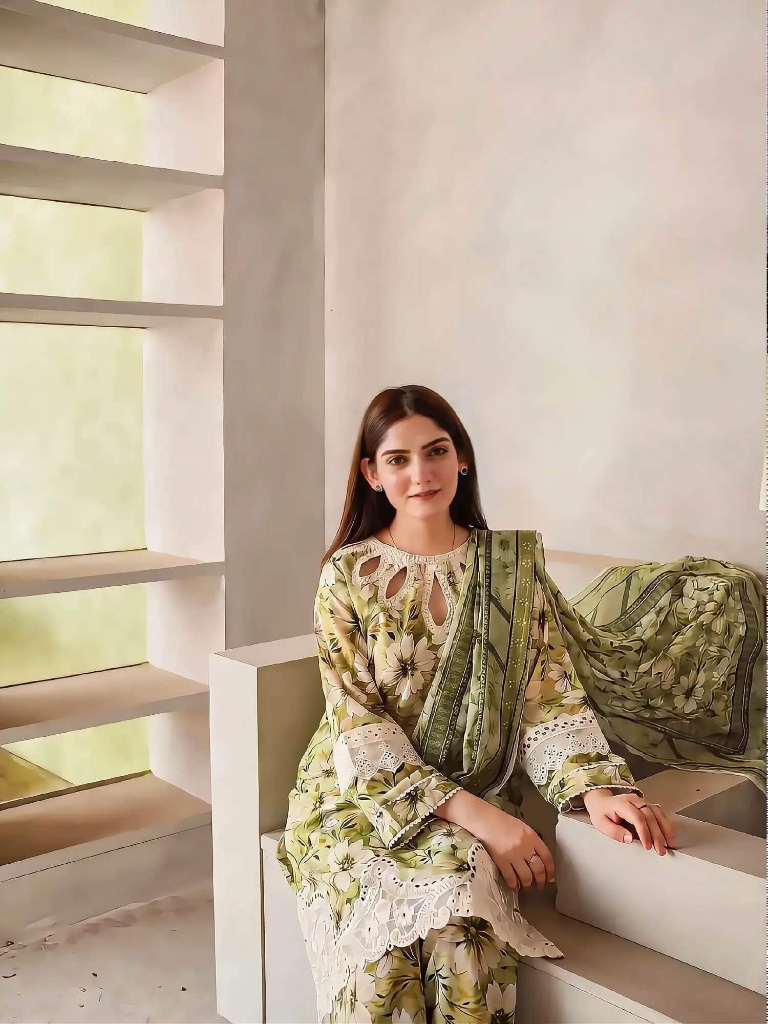 A woman wearing a light green and beige floral printed traditional outfit with white lace trim and a matching dupatta, sitting on a minimalist beige bench in a modern interior setting.