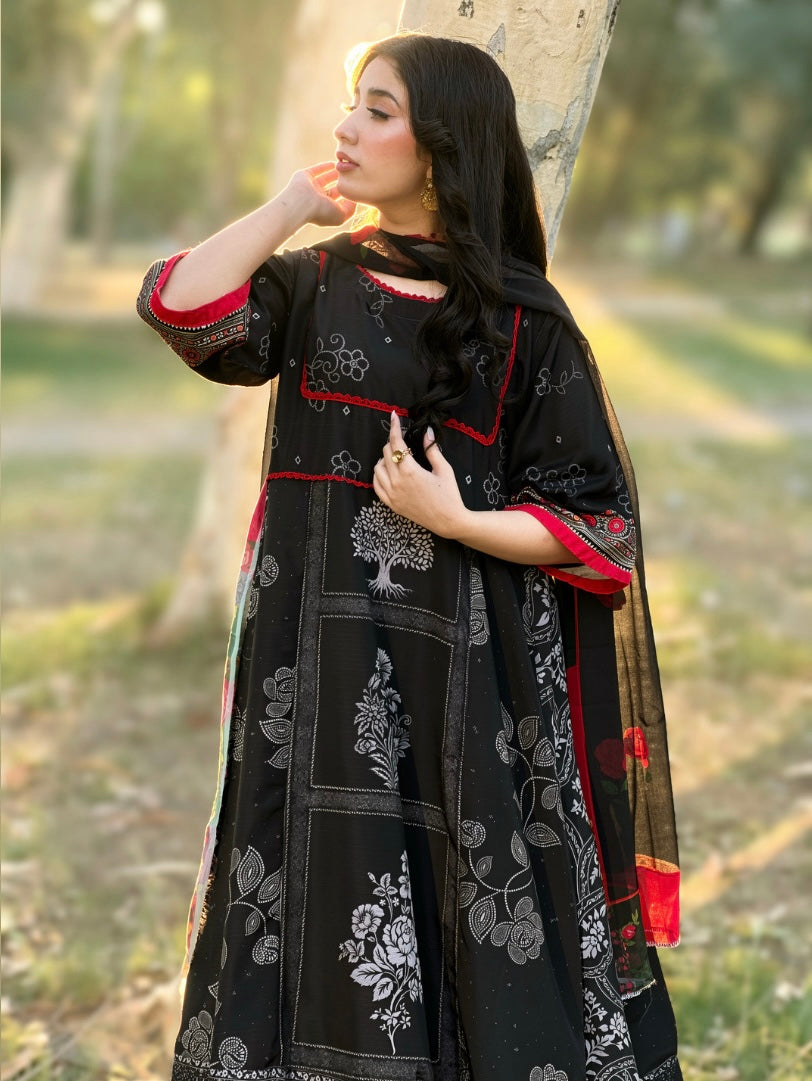 A woman wearing a black traditional outfit with white floral prints and red trim, standing outdoors near a tree in a park-like setting.