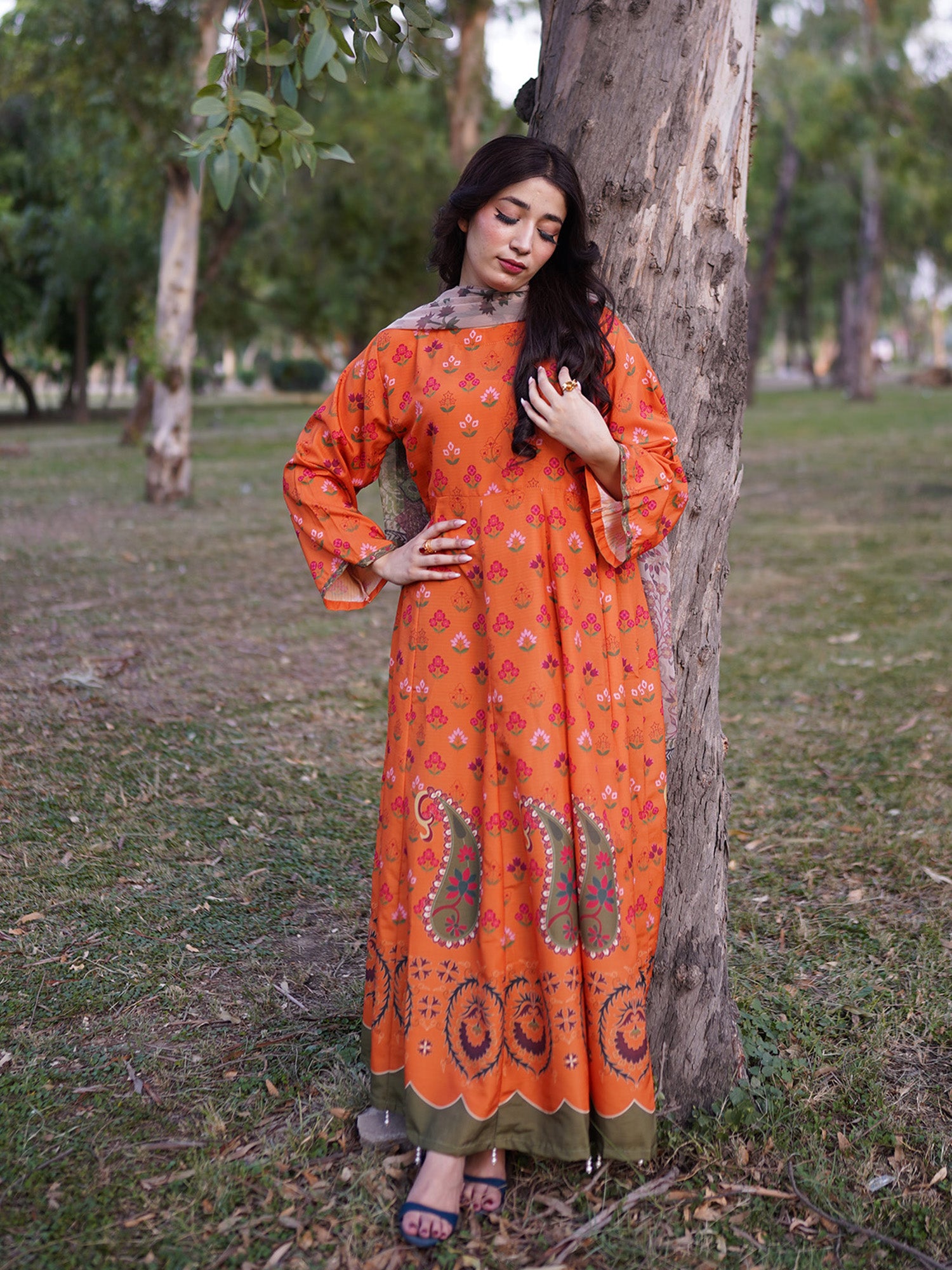 A woman wearing a bright orange long kurta with paisley and floral patterns, standing outdoors next to a tree in a grassy park.