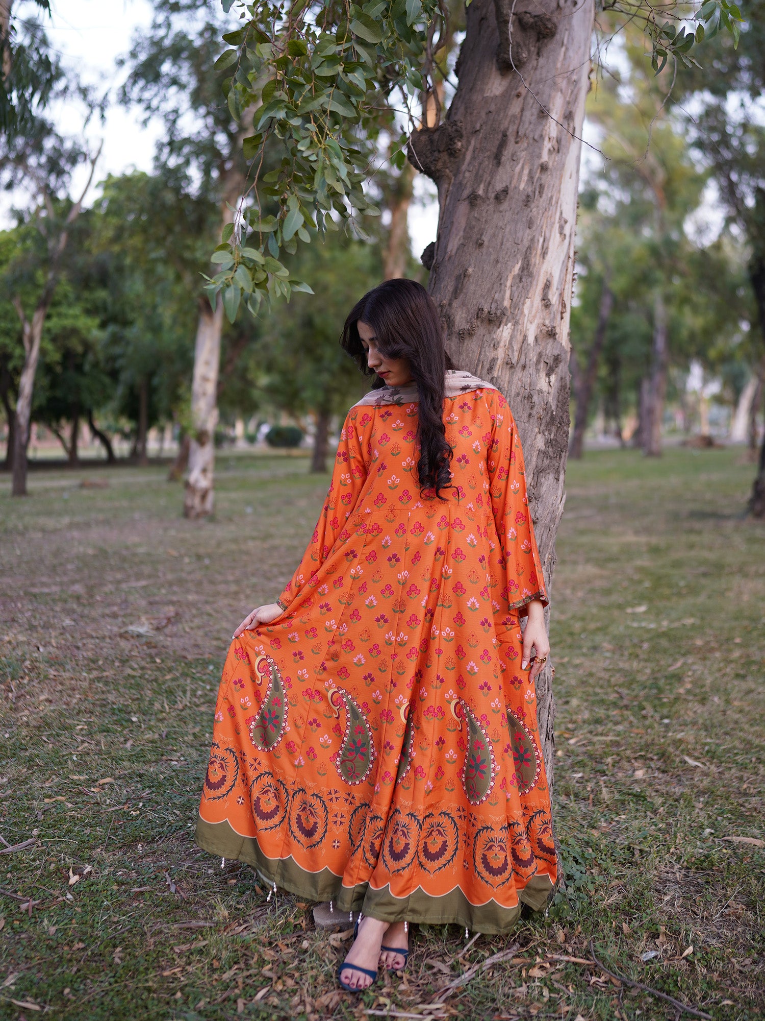 A woman wearing a bright orange printed kurta with paisley and floral motifs, standing outdoors next to a tree in a park.
