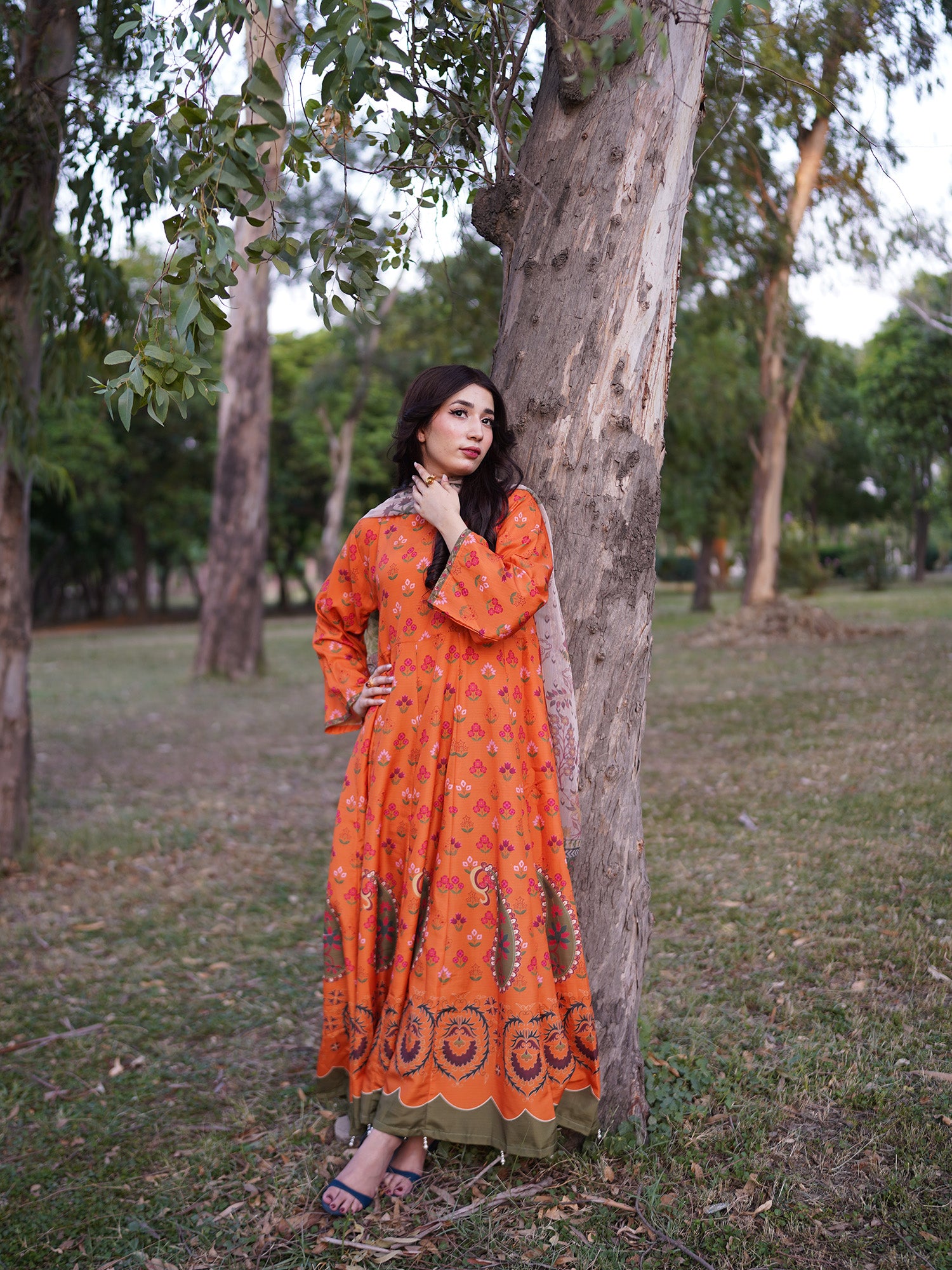 A woman wearing a long orange printed kurta with a green border stands outdoors leaning against a tree in a park setting.