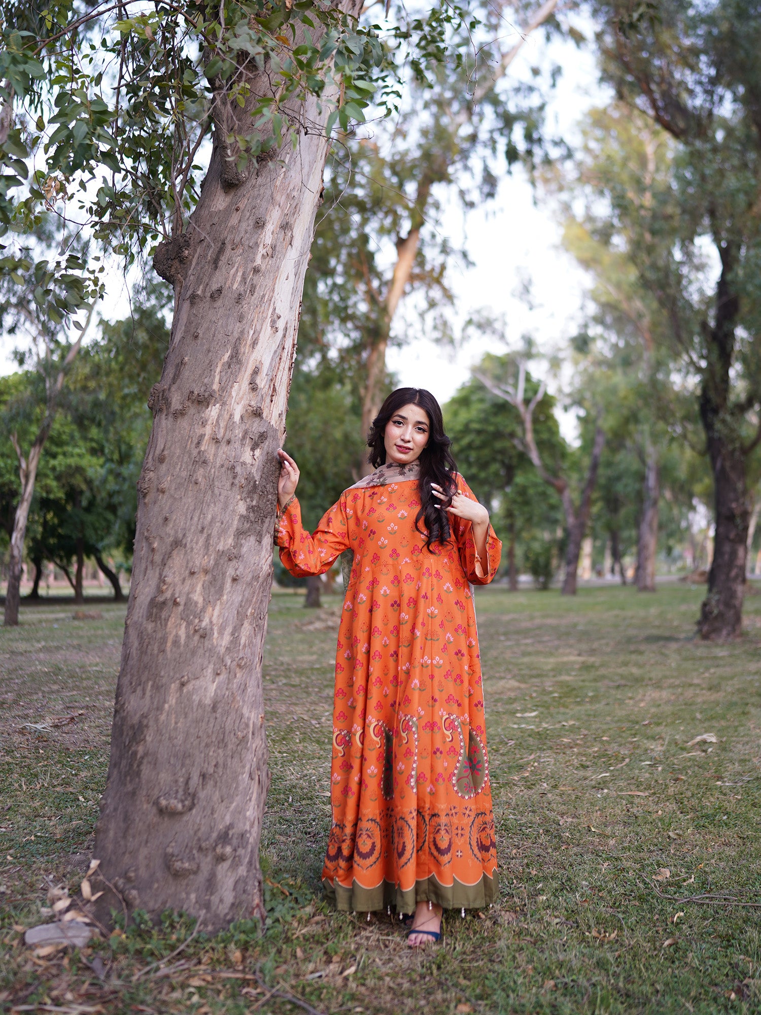 A woman wearing a long orange patterned dress stands outdoors next to a large tree in a park setting.