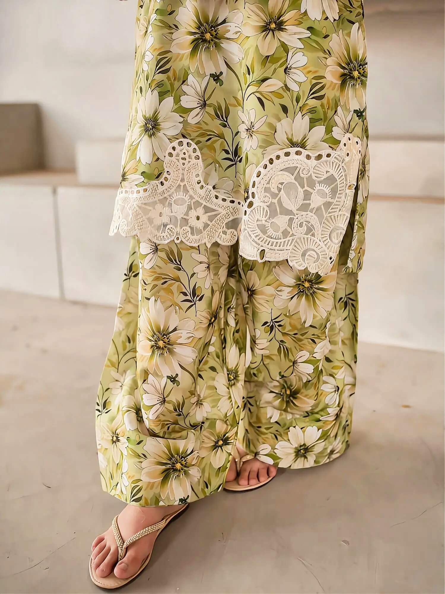A close-up of a person wearing a long, wide-leg maxi skirt with a green and beige floral print and white lace panel details, paired with beige thong sandals.