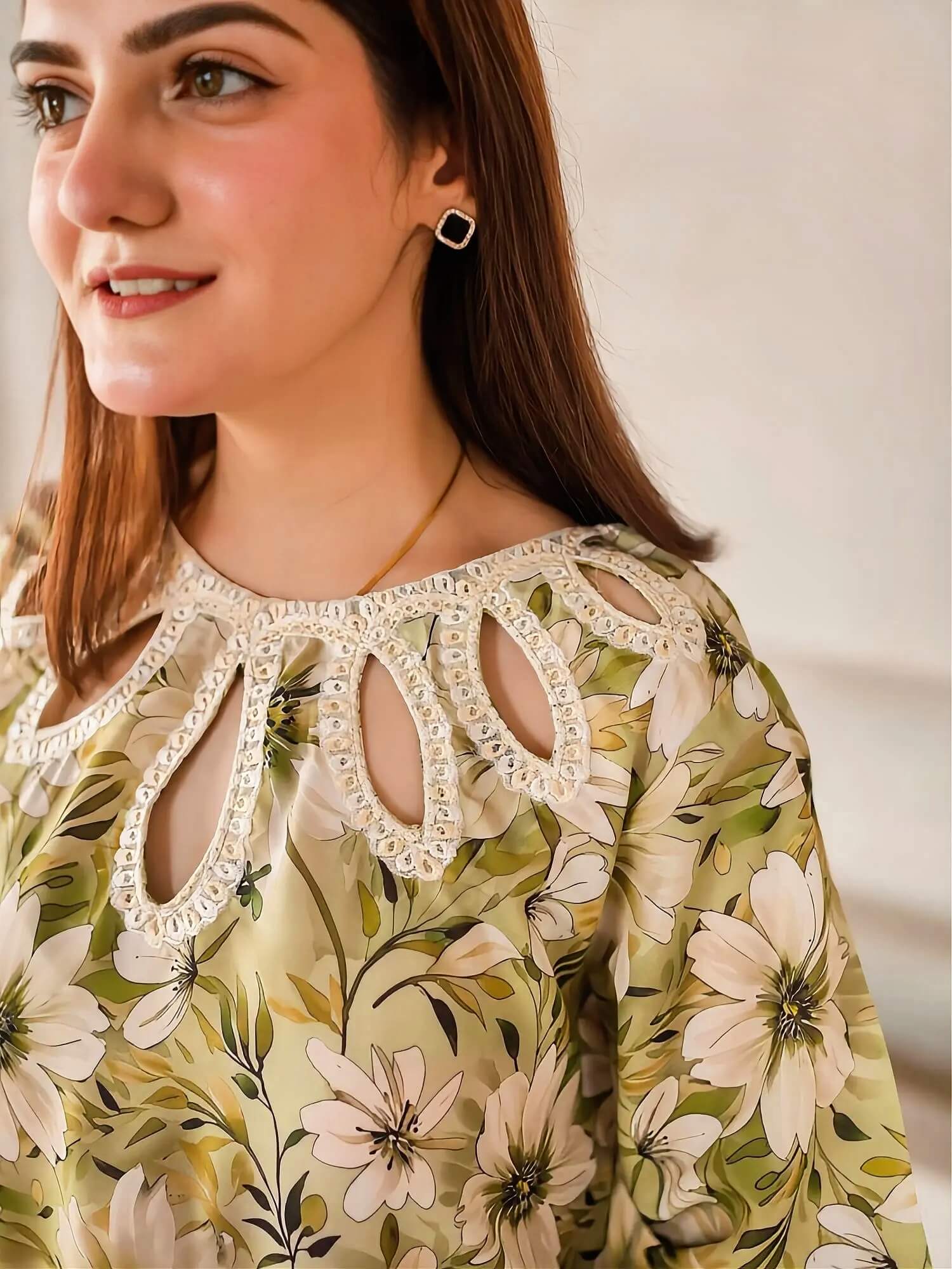 A close-up of a woman wearing a floral long-sleeve top with a decorative cutout neckline and a cream lace trim. The top has a green, beige, and white floral pattern.