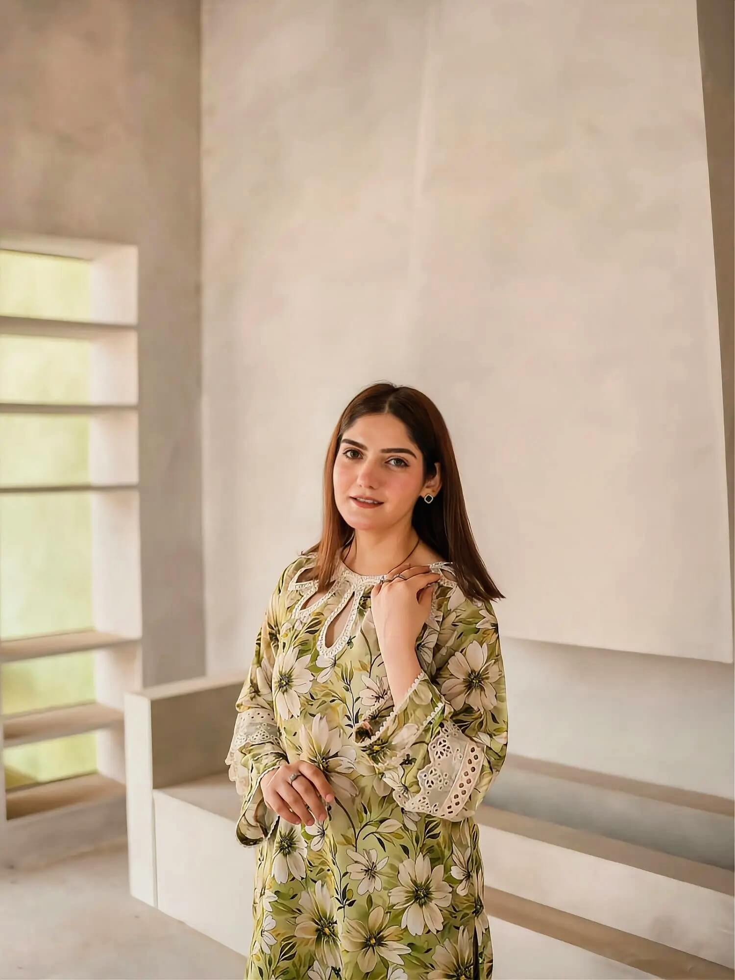 A woman wearing a green and beige floral print tunic dress with lace sleeves, standing in a minimalist, modern interior with concrete walls and built-in shelves.