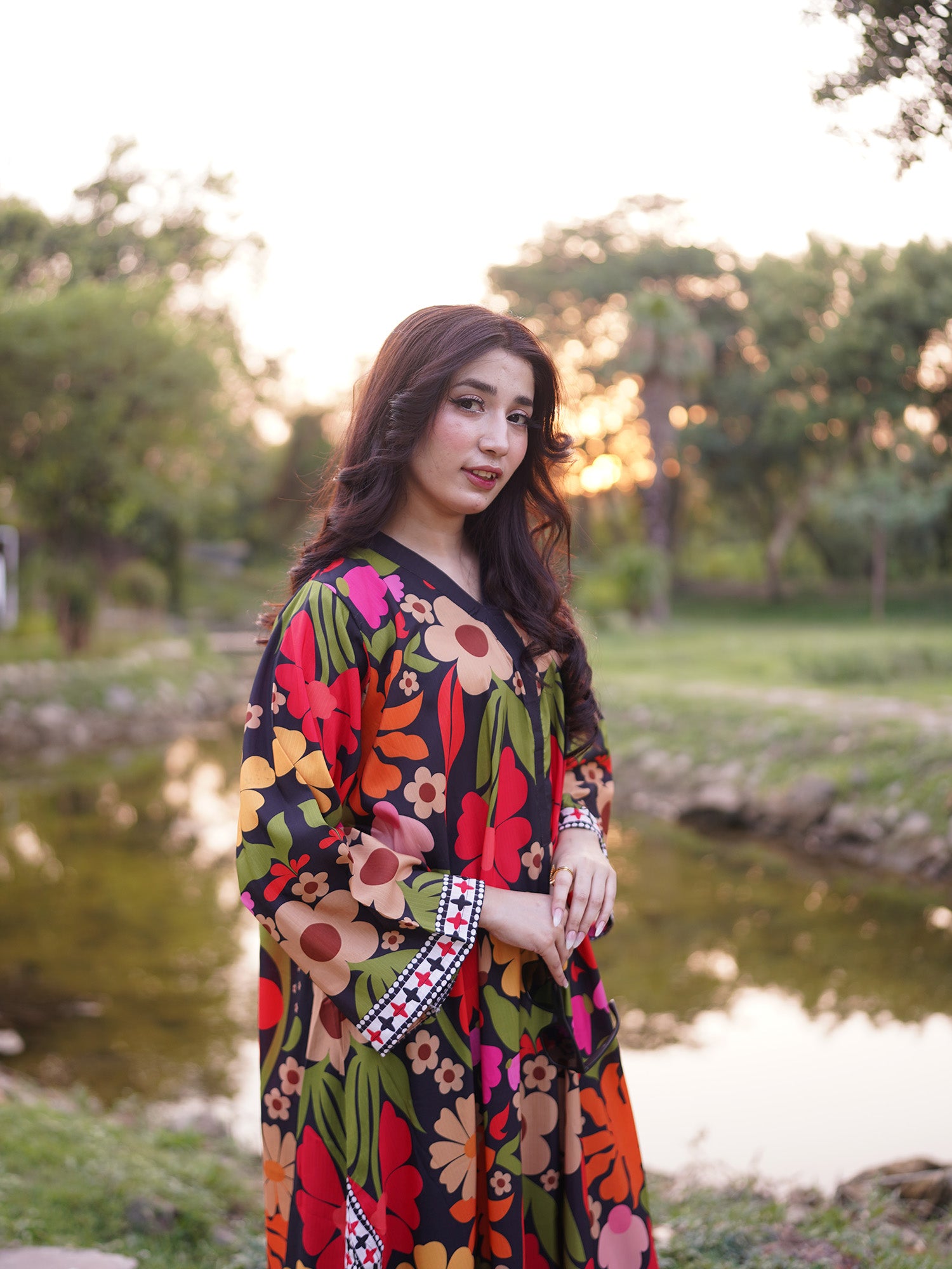 A woman wearing a long-sleeved black kaftan dress with a vibrant multicolor floral print stands outdoors near a pond during sunset.