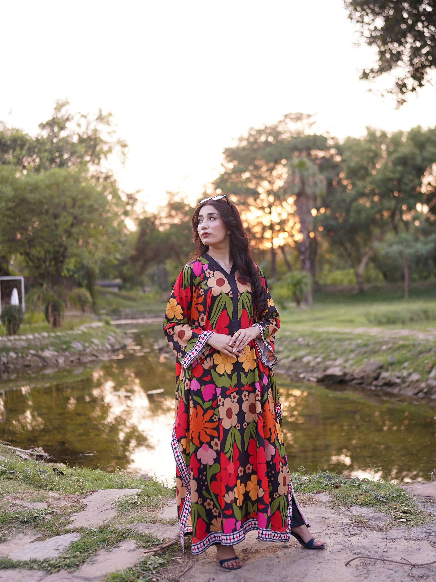 A woman wearing a long-sleeved, floor-length floral print kaftan dress stands outdoors next to a pond in a park during sunset. The dress features a vibrant multicolor floral pattern with red, orange, yellow, pink, and green on a black base, with patterned borders.