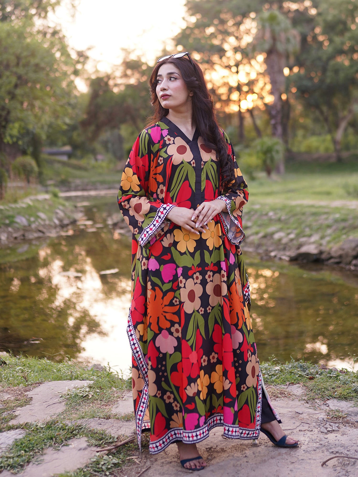 A woman wearing a long-sleeved black maxi dress with a vibrant multicolor floral print stands outdoors near a pond at sunset.