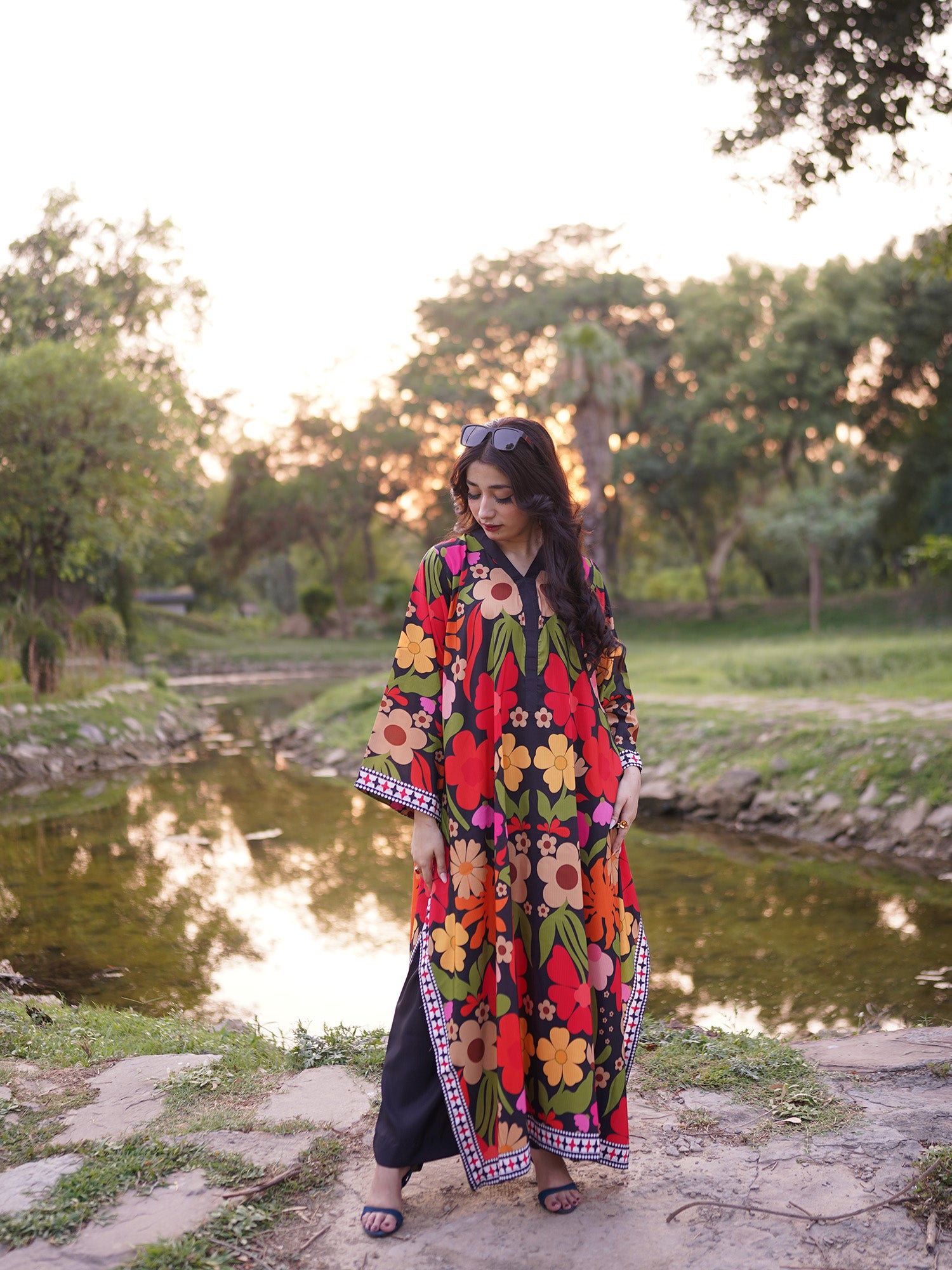 A woman stands outdoors by a small pond at sunset wearing a long, loose floral kaftan dress with wide sleeves and black wide-leg pants underneath. The dress has a bold multicolor floral print on a black base with red, yellow, orange, green, and pink flowers.