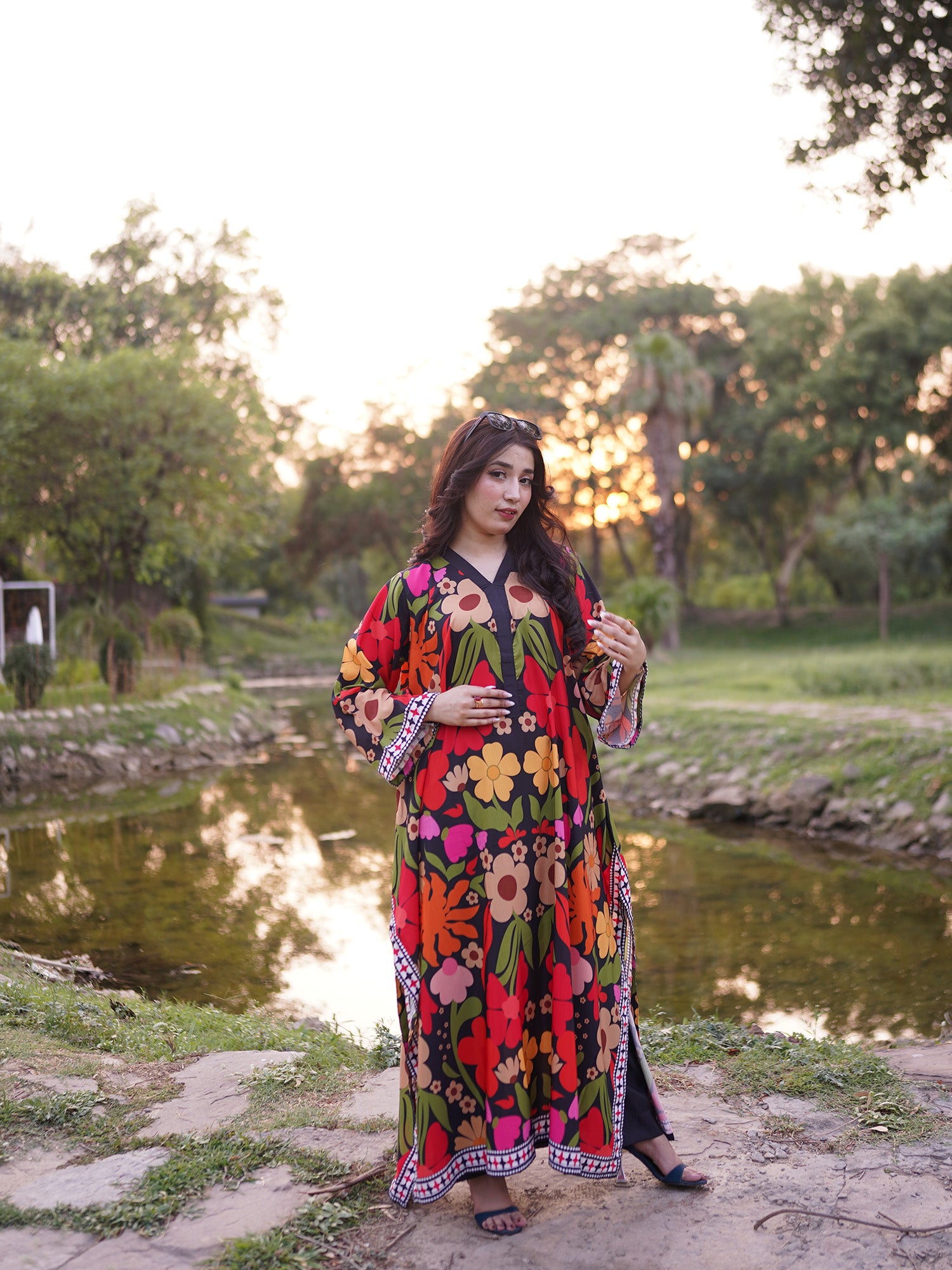A woman wearing a vibrant, multicolored floral print kaftan dress stands outdoors next to a pond during sunset, with trees in the background.