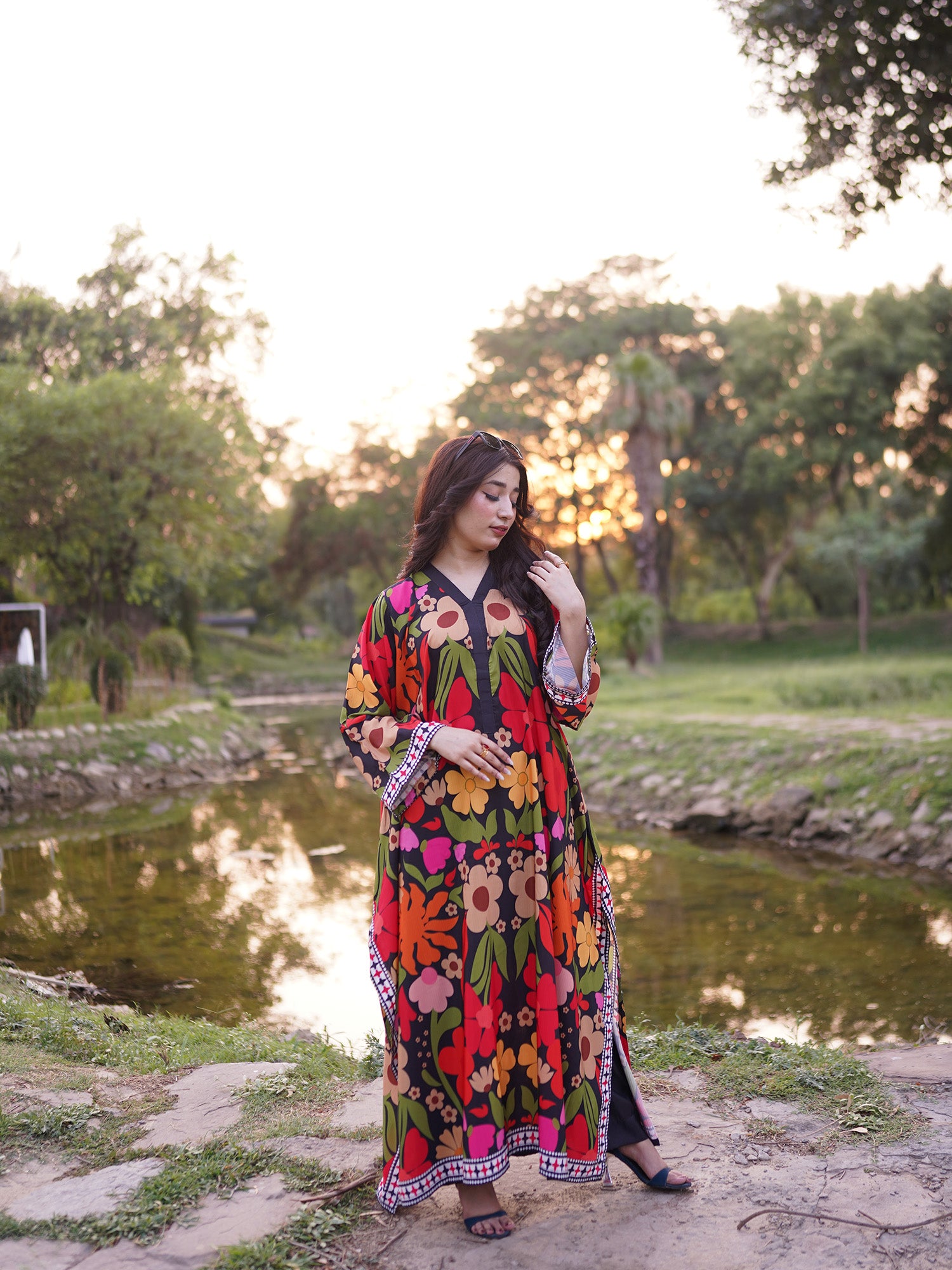 A woman wearing a vibrant, multicolored floral print maxi dress stands outdoors by a pond at sunset. The dress features a bold pattern of red, yellow, orange, green, and pink flowers on a black background, with a patterned border.