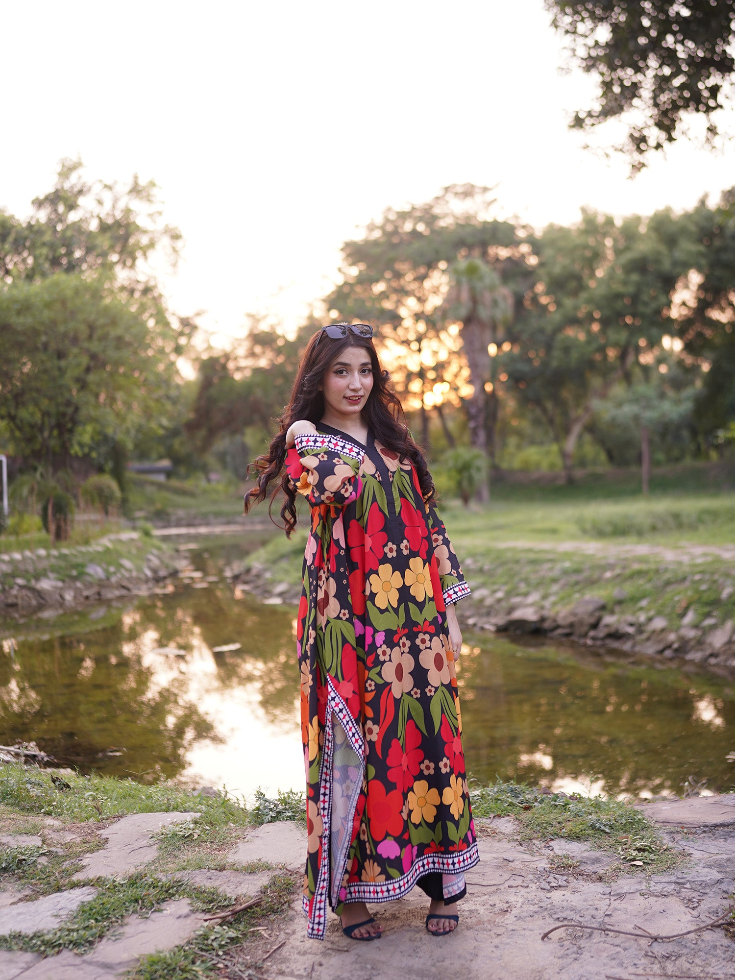A woman stands outdoors beside a small pond in a park, wearing a long, off-the-shoulder floral dress with a multicolor print of red, yellow, green, and orange flowers on a dark background.