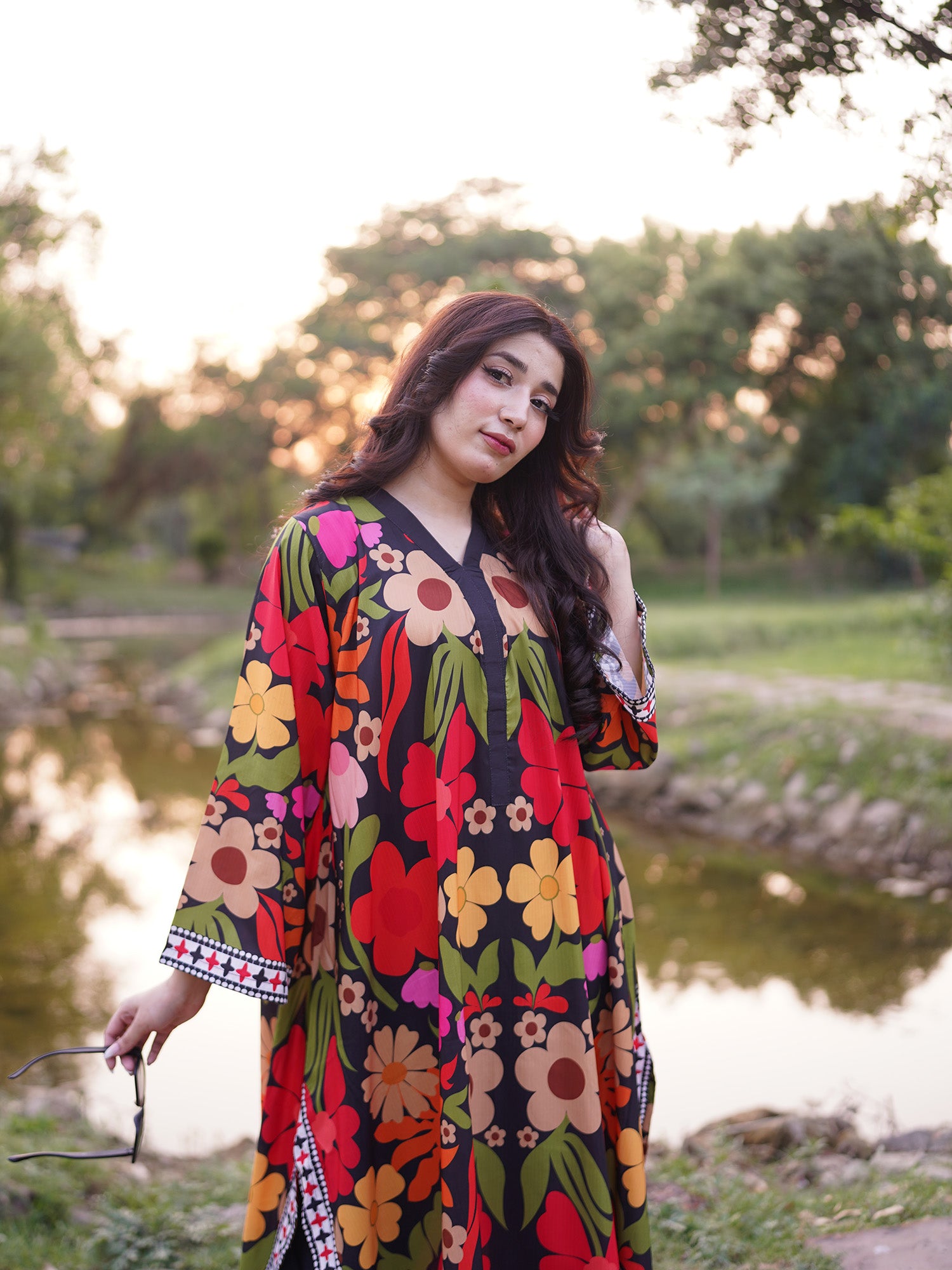 A woman wearing a long-sleeved black kaftan dress with a vibrant multicolor floral print stands outdoors near a pond during sunset.