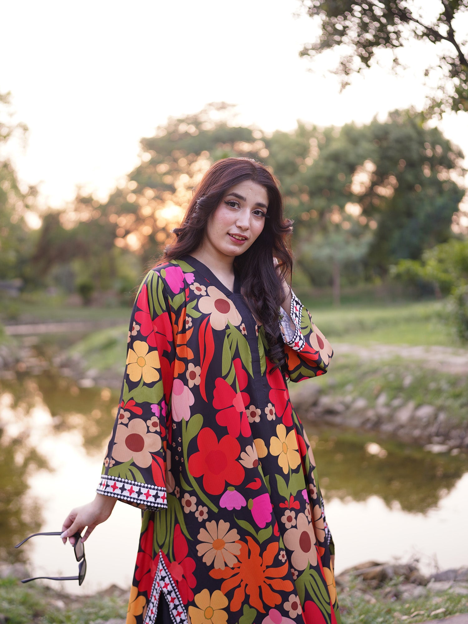 A woman wearing a vibrant, multicolored floral print kaftan dress stands outdoors near a pond during sunset. The dress features a black base with large red, pink, orange, yellow, and green flowers.