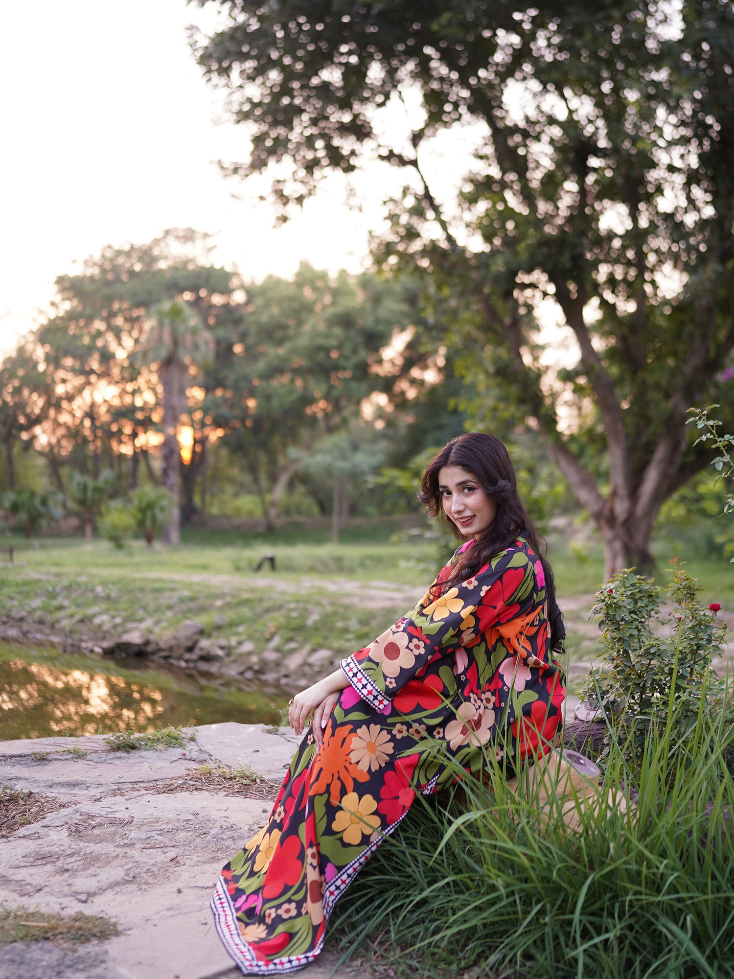 A woman wearing a vibrant, multicolored floral printed kaftan sits on a stone path in a park setting during golden hour, with trees and grass in the background.
