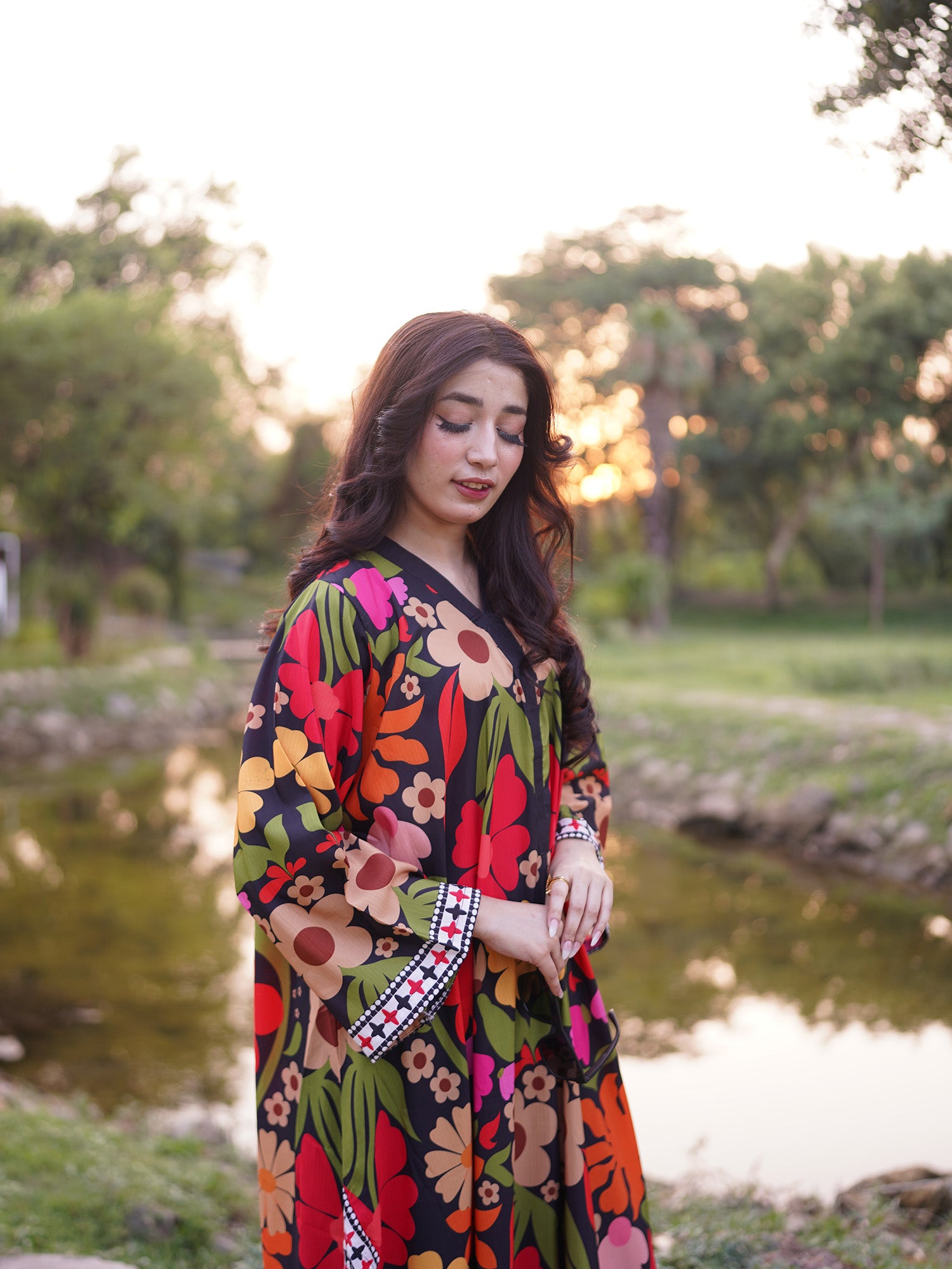 A woman wearing a vibrant, multicolored floral print kaftan dress stands outdoors near a pond during sunset, with trees in the background.