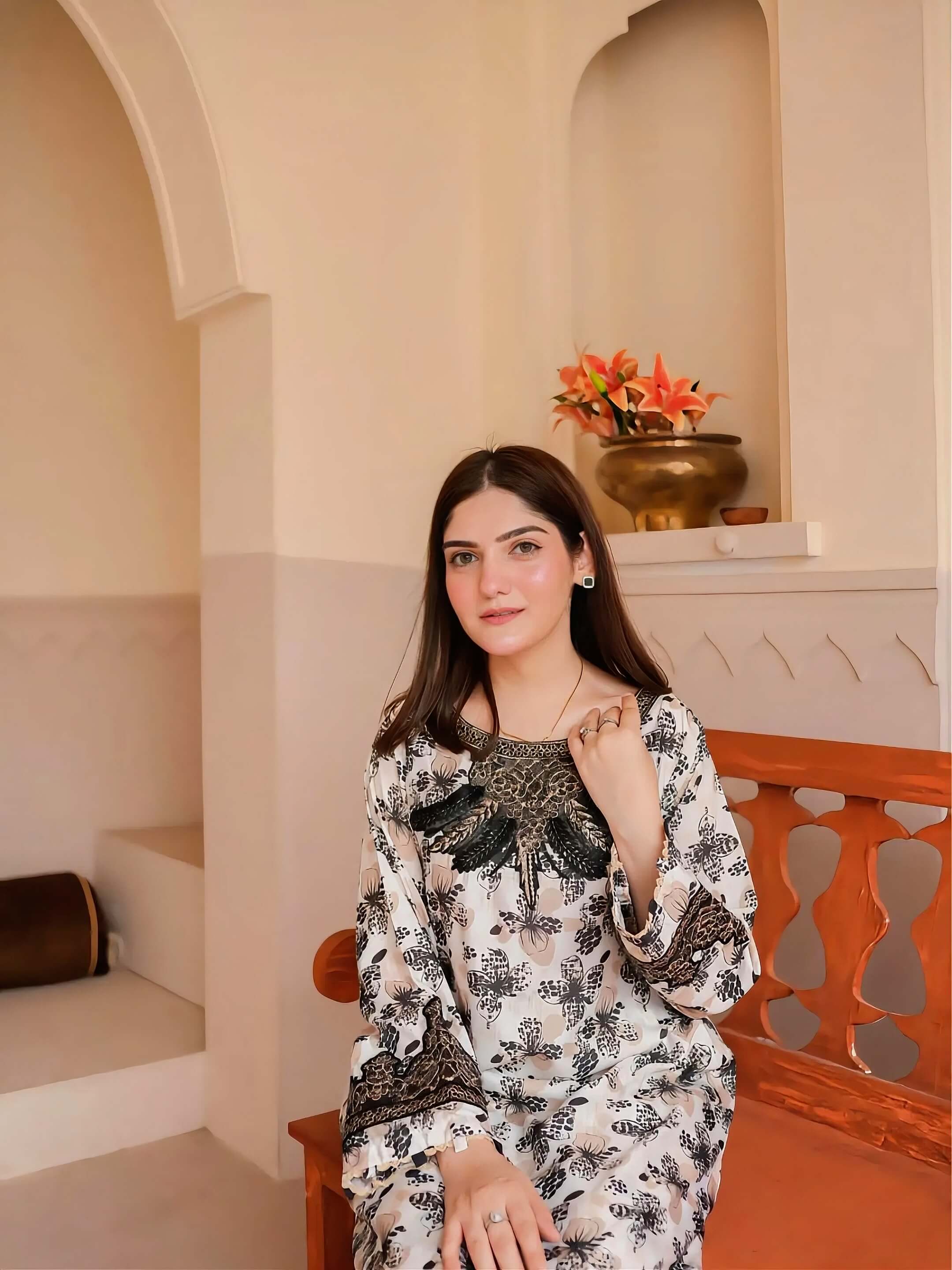 A woman wearing a white long-sleeve kurta with black floral patterns and a black embroidered yoke sits on a wooden bench in a warm, arched interior setting.