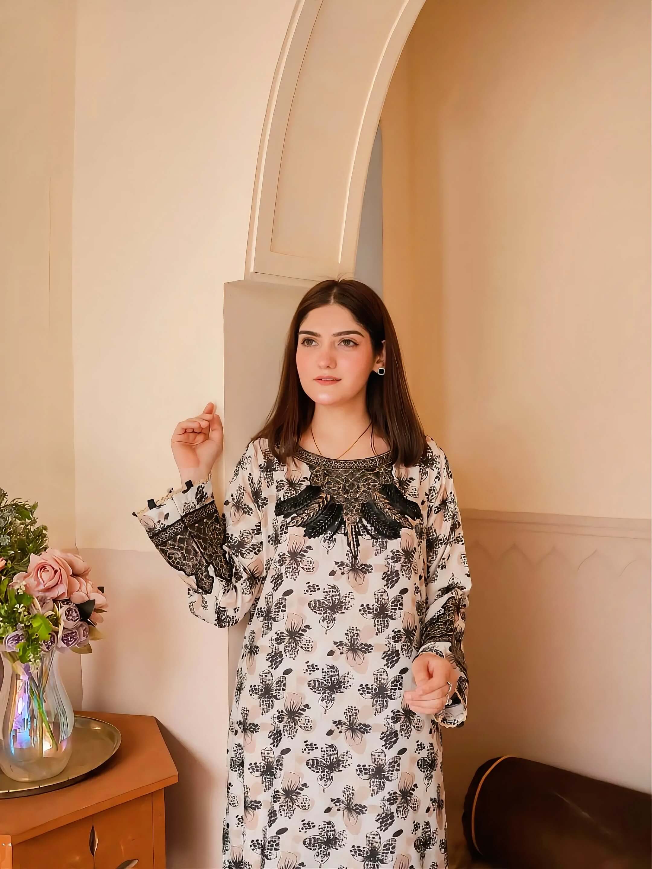 A woman wearing a long-sleeved cream-colored dress with a black floral pattern and black lace detailing on the chest and cuffs, standing in an indoor setting with beige walls and an arched doorway.
