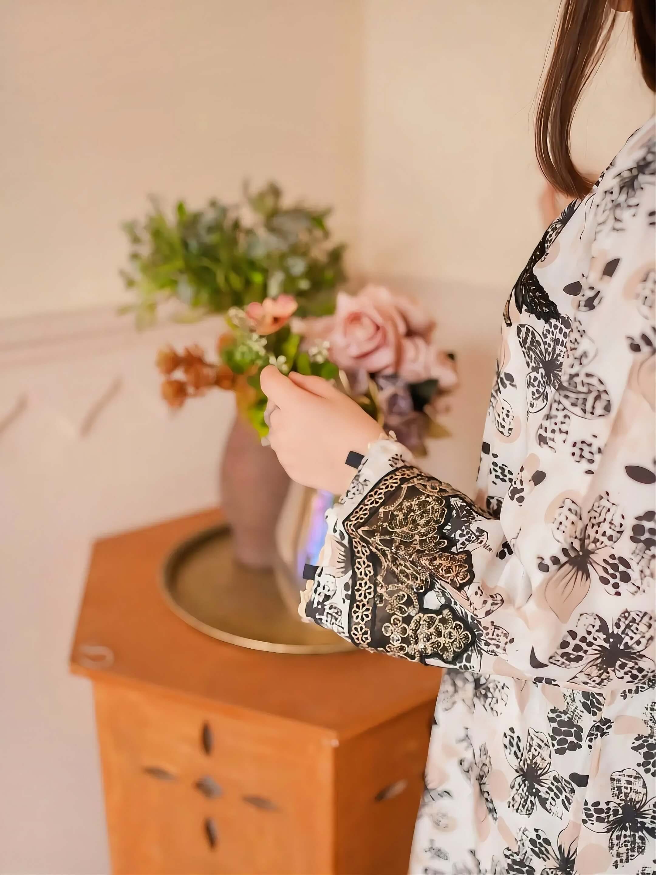 A woman wearing a white long-sleeve blouse with a black and beige floral and paisley print is shown from the side, holding a bouquet of pink and green flowers on a wooden side table.