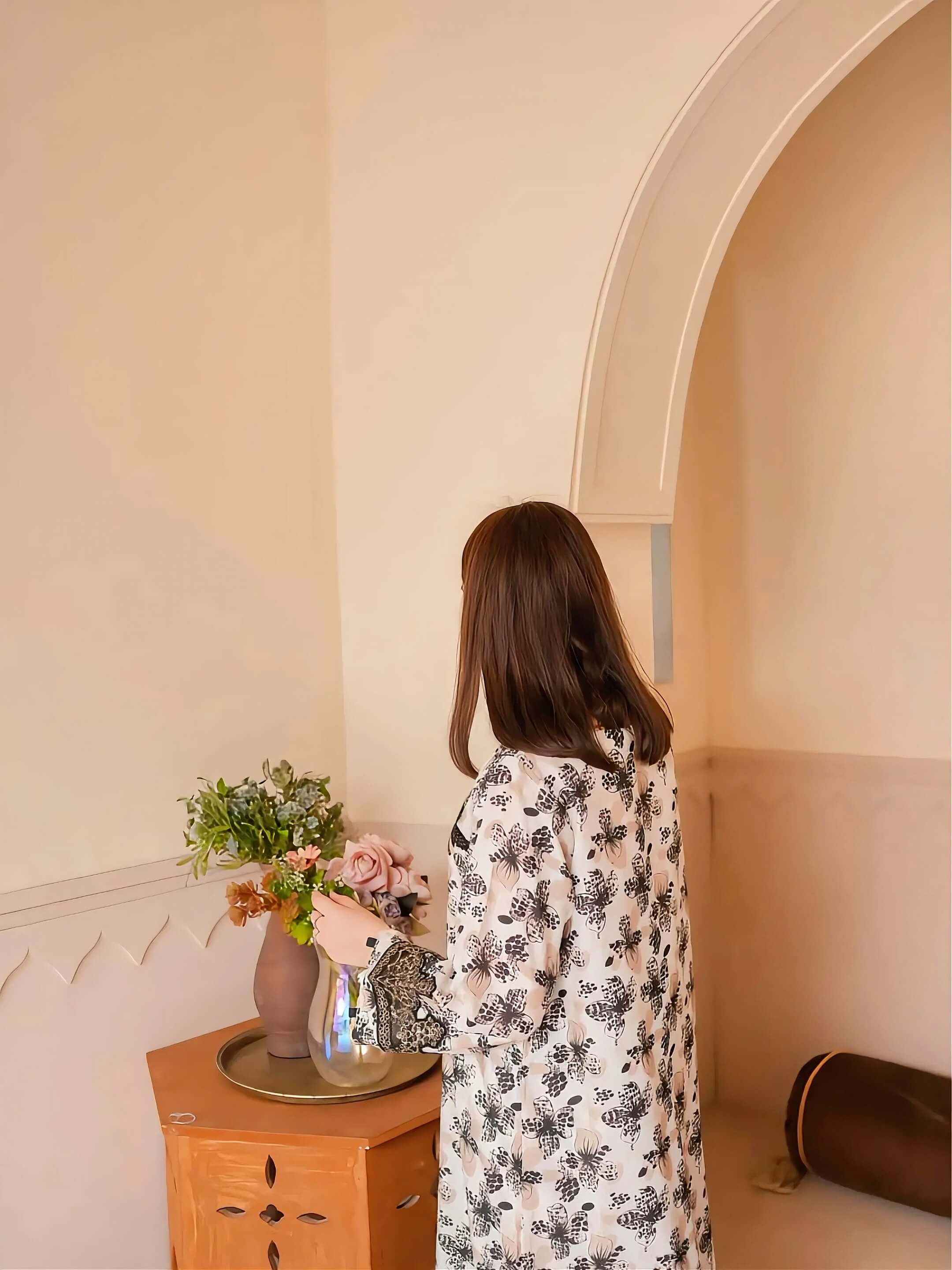 A woman with long brown hair stands with her back to the camera, wearing a white long-sleeve button-up shirt with a black floral pattern. She is holding a glass vase filled with pink and green flowers on a small wooden side table in a warm, beige-toned room with an arched doorway.