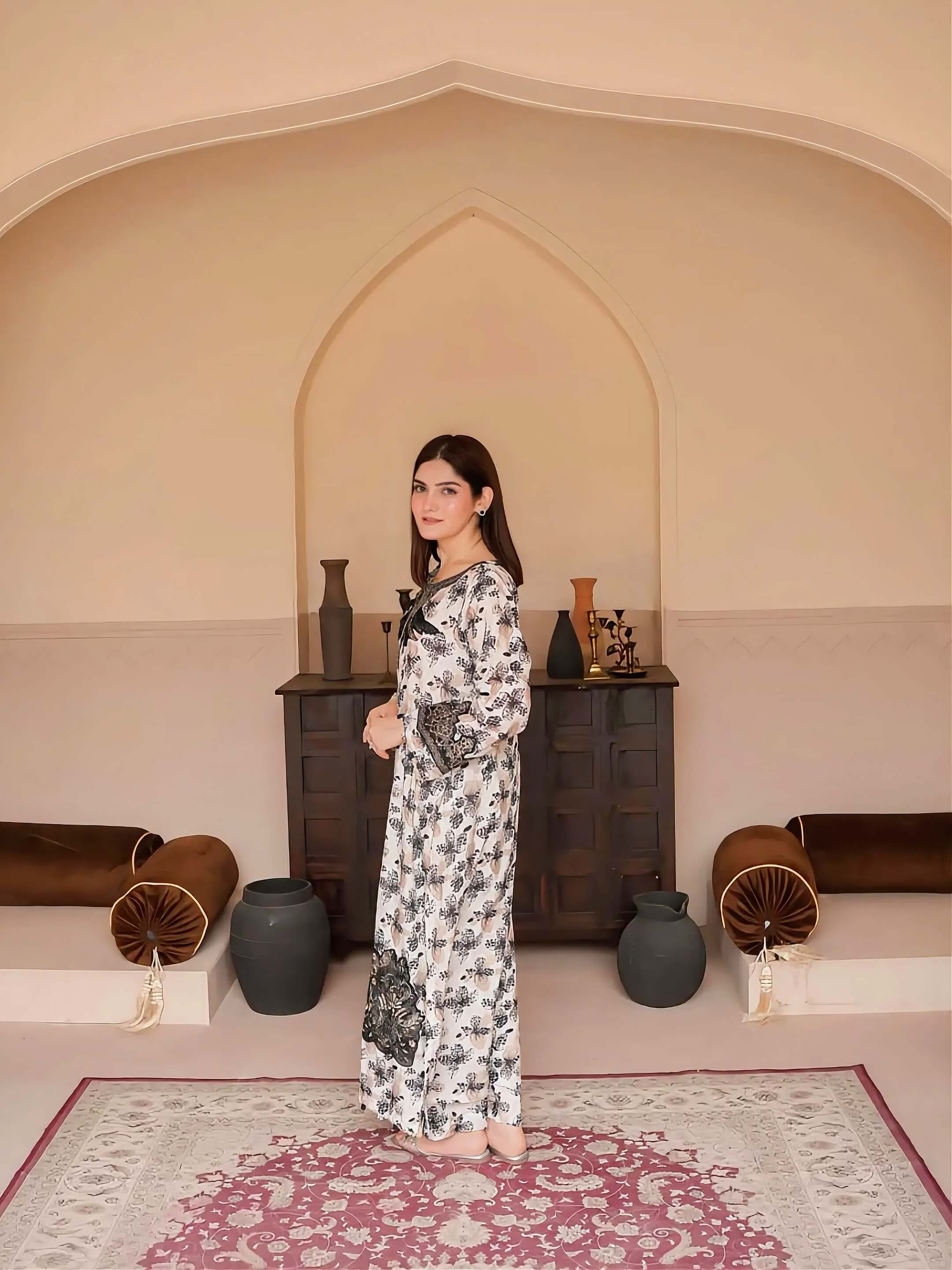 A woman stands in a decorated room with arched wall niches, wearing a long white maxi dress with a black floral print. She is positioned in front of a dark wooden cabinet with decorative vases, with cushions and a patterned rug on the floor.
