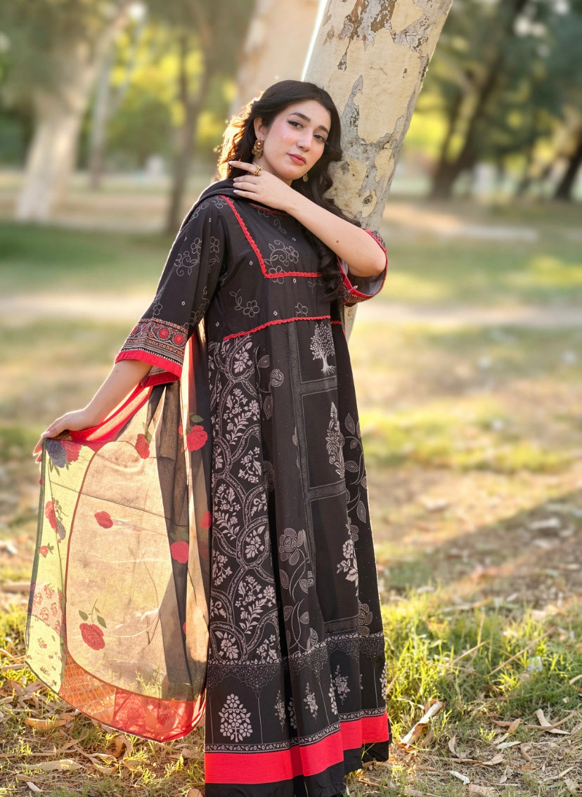 A woman wearing a black printed Anarkali kurta with red border accents and a beige dupatta with red motifs, posing outdoors next to a tree in a park.