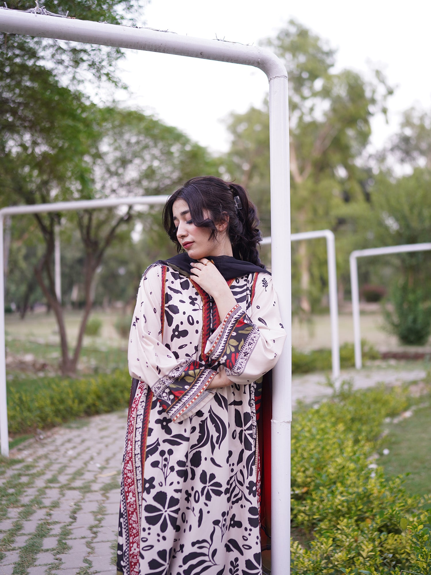 A woman wearing a traditional long-sleeve embroidered dress with black floral patterns on a cream base, standing outdoors next to a white metal frame in a park-like setting.
