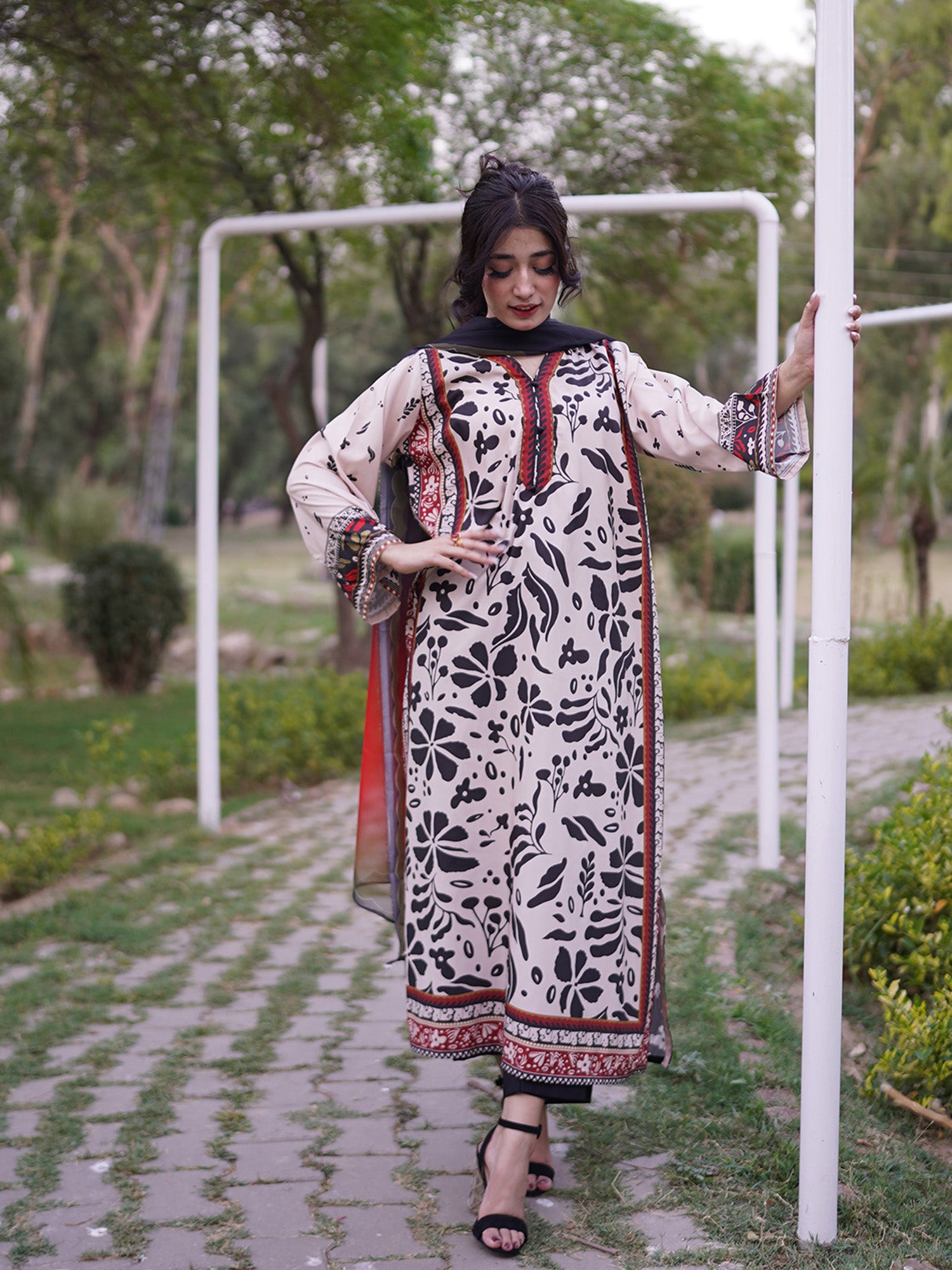 A woman wearing a cream-colored traditional kurta with black floral patterns and red border details, standing outdoors on a paved path in a park-like setting.