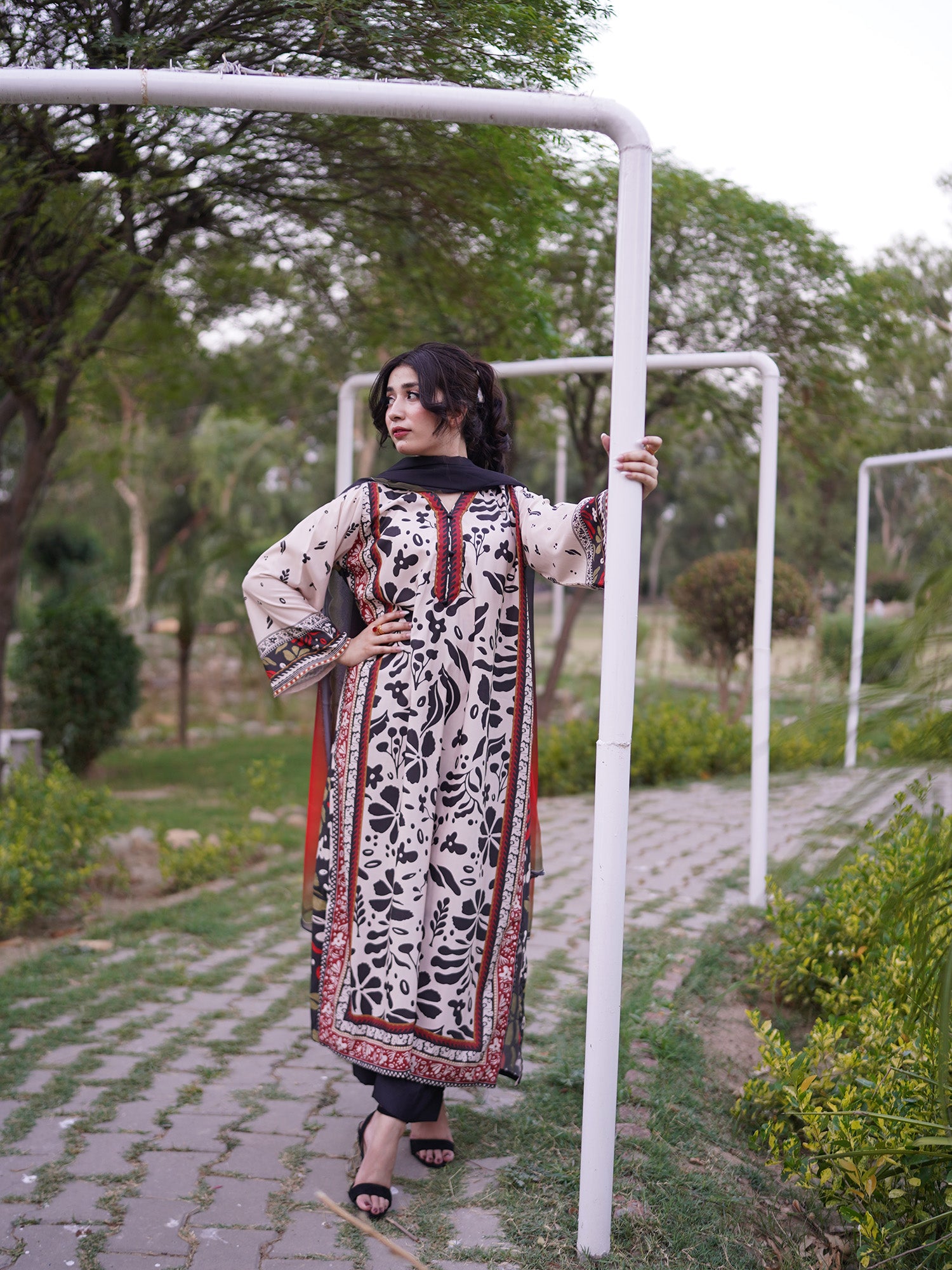 A woman stands outdoors on a paved path wearing a long cream-colored printed kurta with black floral motifs and red border details, posing next to a white metal frame in a park-like setting.