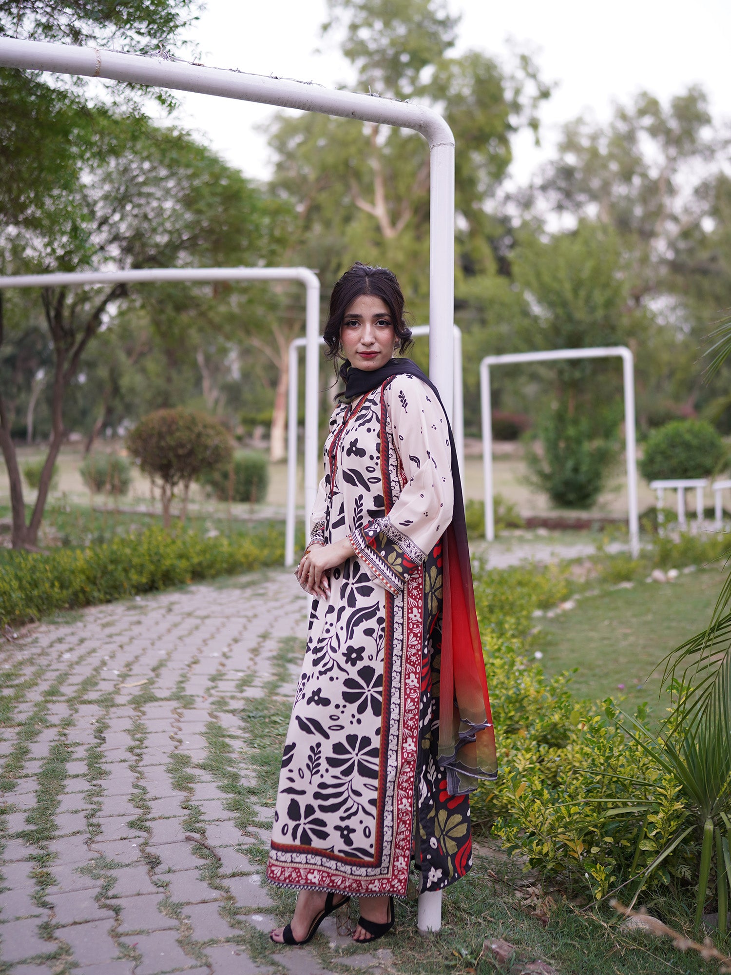 A woman stands outdoors on a paved path wearing a long cream-colored traditional outfit with black floral patterns and a red border, paired with a red and black dupatta. The background shows a park with green trees and white metal frames.