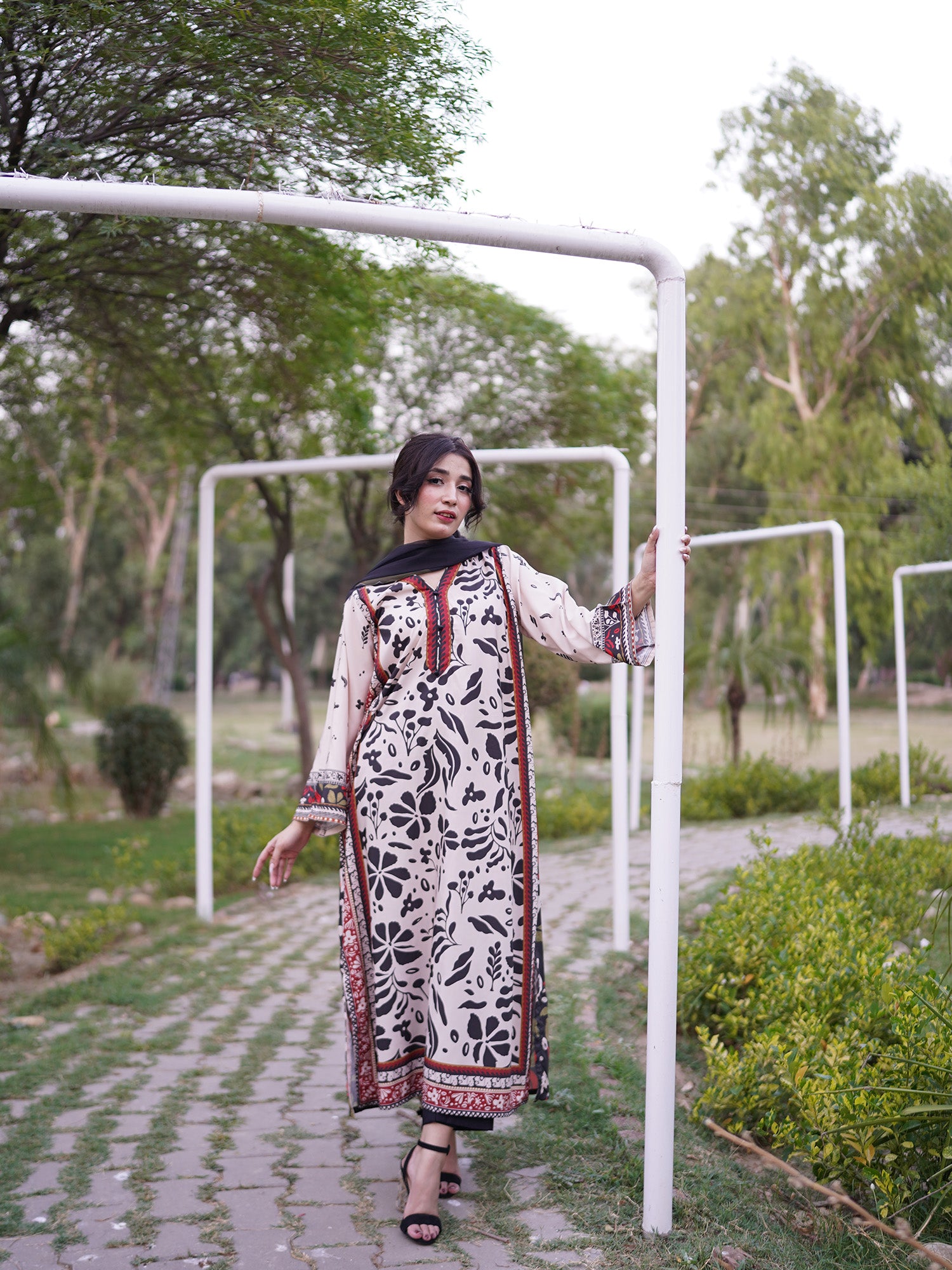 A woman wearing a long black and white patterned kurta with red border details walks along a paved path in a park, holding onto a white metal frame.