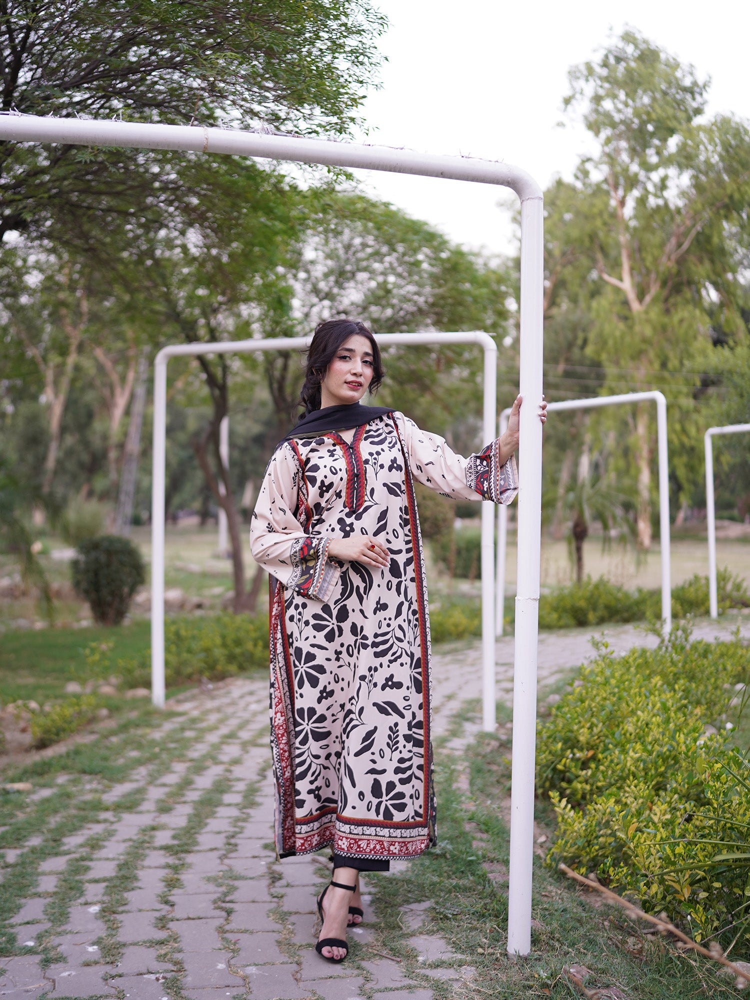 A woman wearing a long-sleeved beige and black floral print kurta with red patterned borders and a black dupatta, standing in a park next to a white metal frame.