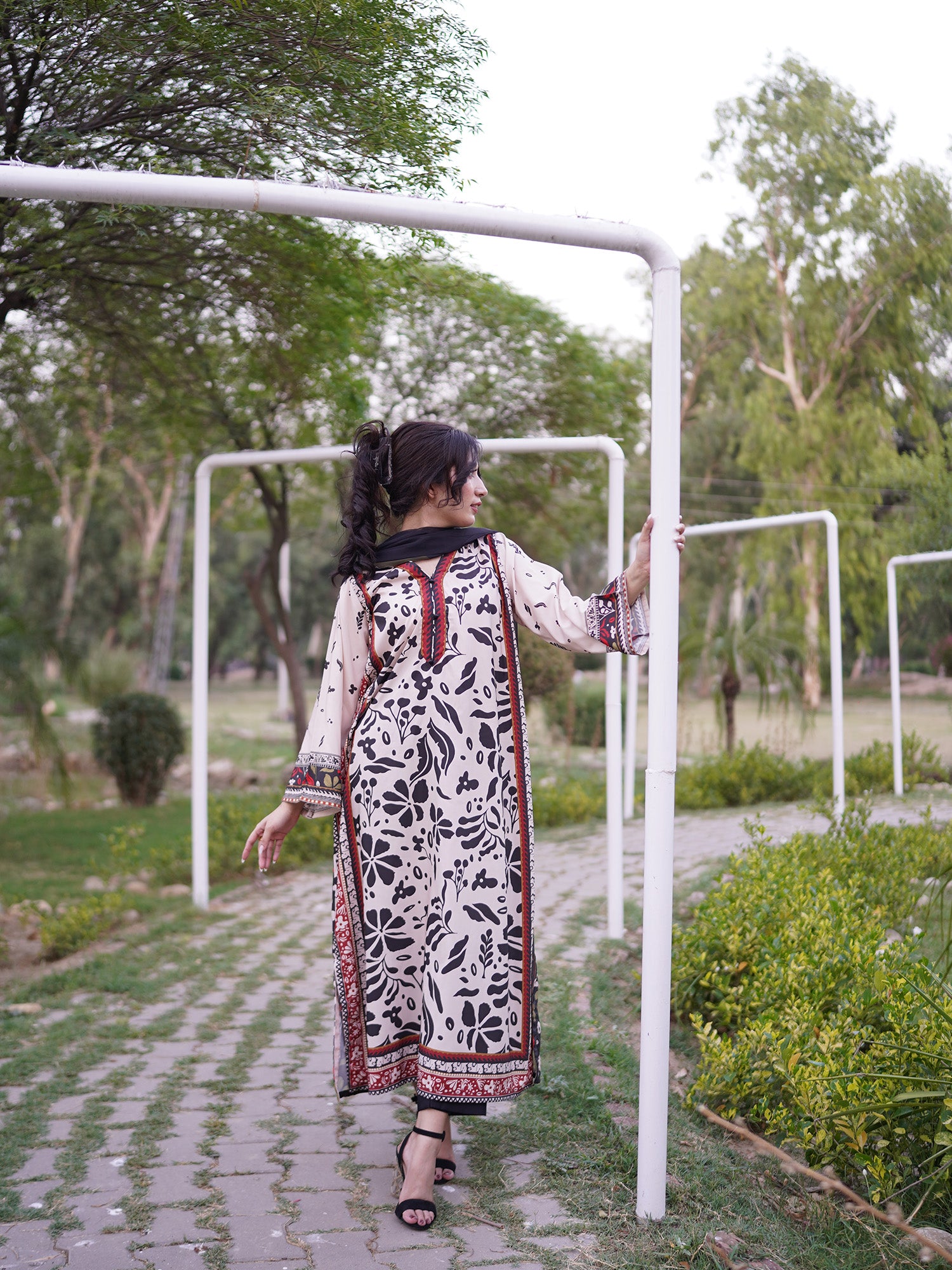 A woman wearing a long white kurta with black floral print and red border details walks along a paved path in a park, holding onto a white metal frame.