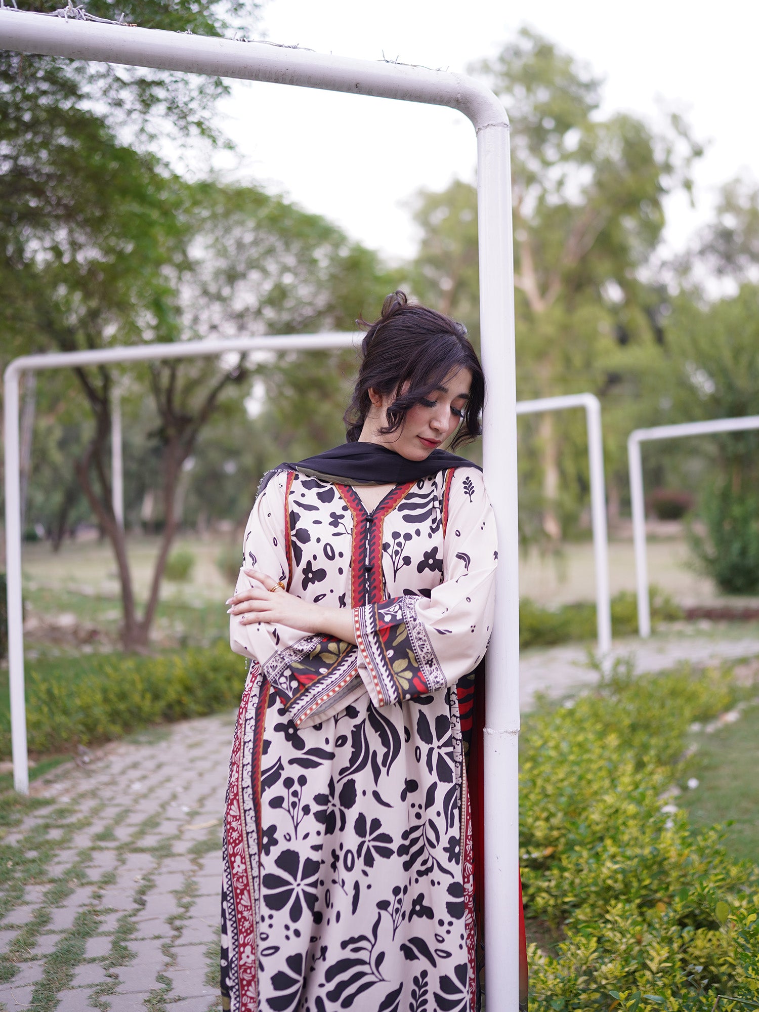 A woman wearing a traditional patterned cream and black kurta with red and green border details, standing outdoors in a park-like setting next to a white metal frame.